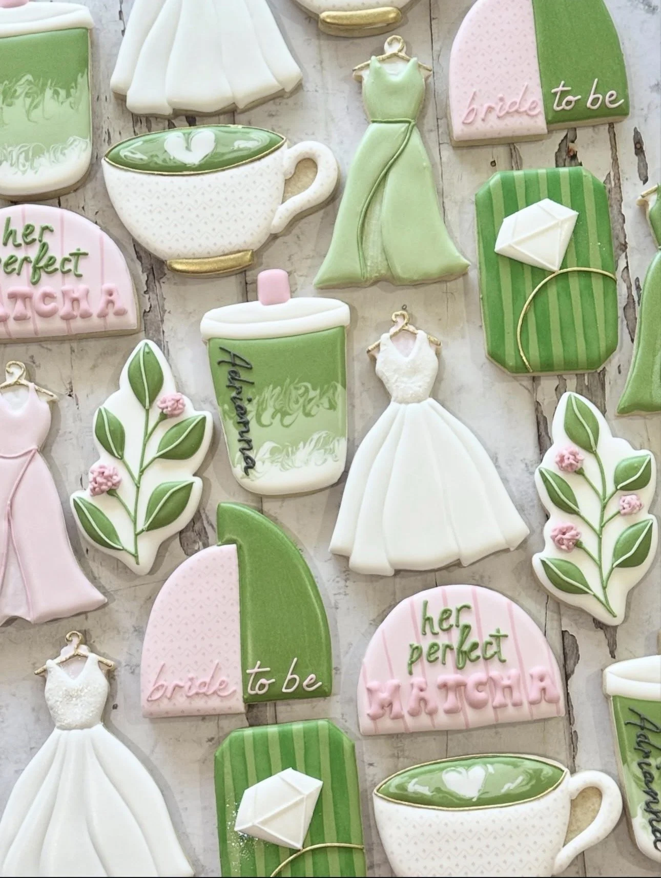 Assorted bridal shower cookies decorated as wedding dresses, rings, teacups, leaves, presents, and green-and-pink designs reading “bride to be” and “her perfect match.”