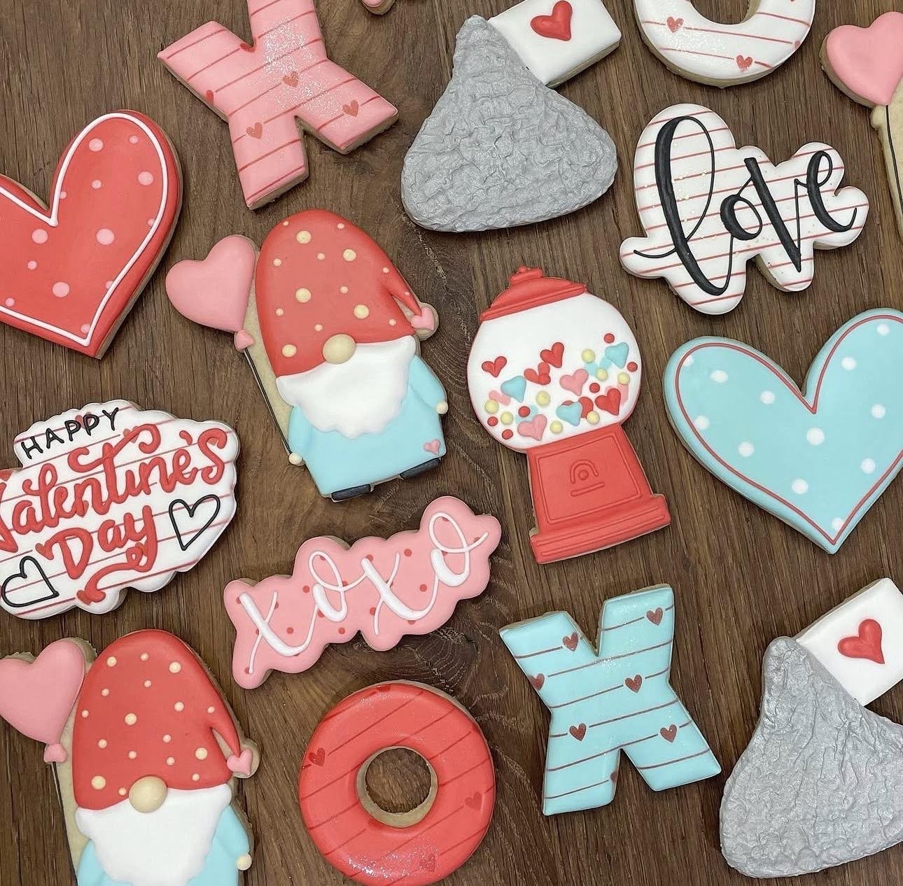 Colorful Valentine's Day decorated sugar cookies, including hearts, gnomes, a candy dispenser, and love-themed messages on a wooden table