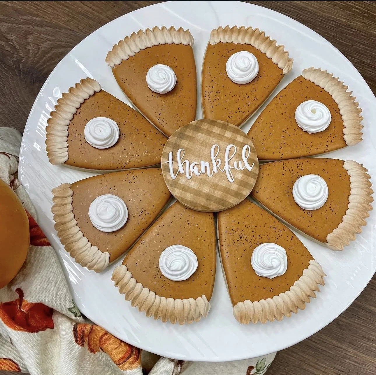 Pumpkin pie slices arranged in a circle on a white platter, topped with whipped cream and a center plaque reading “thankful.”