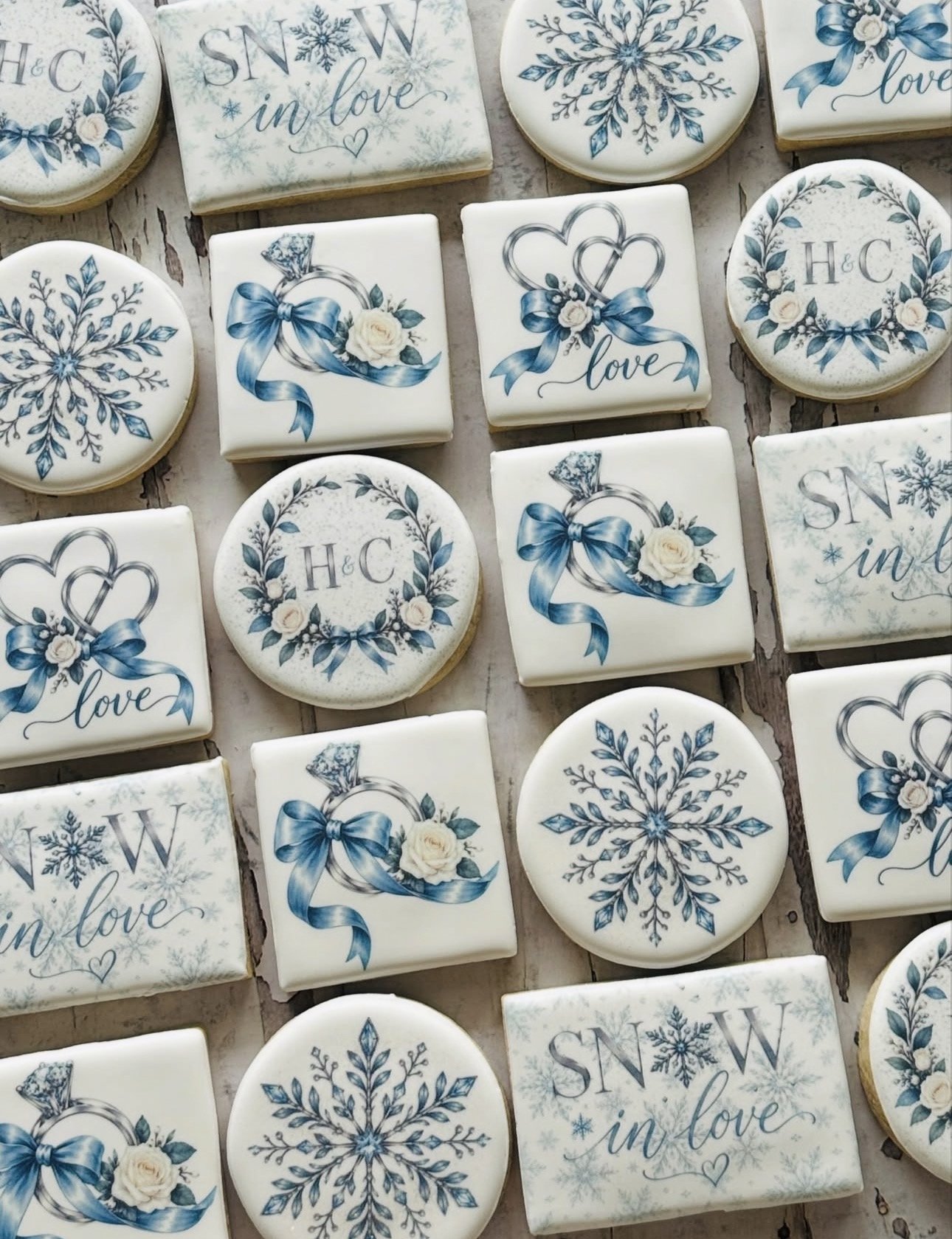 Assorted decorative wedding cookies with blue ribbons, white roses, snowflakes, hearts, and “SNOW in love” and “H&C” designs.