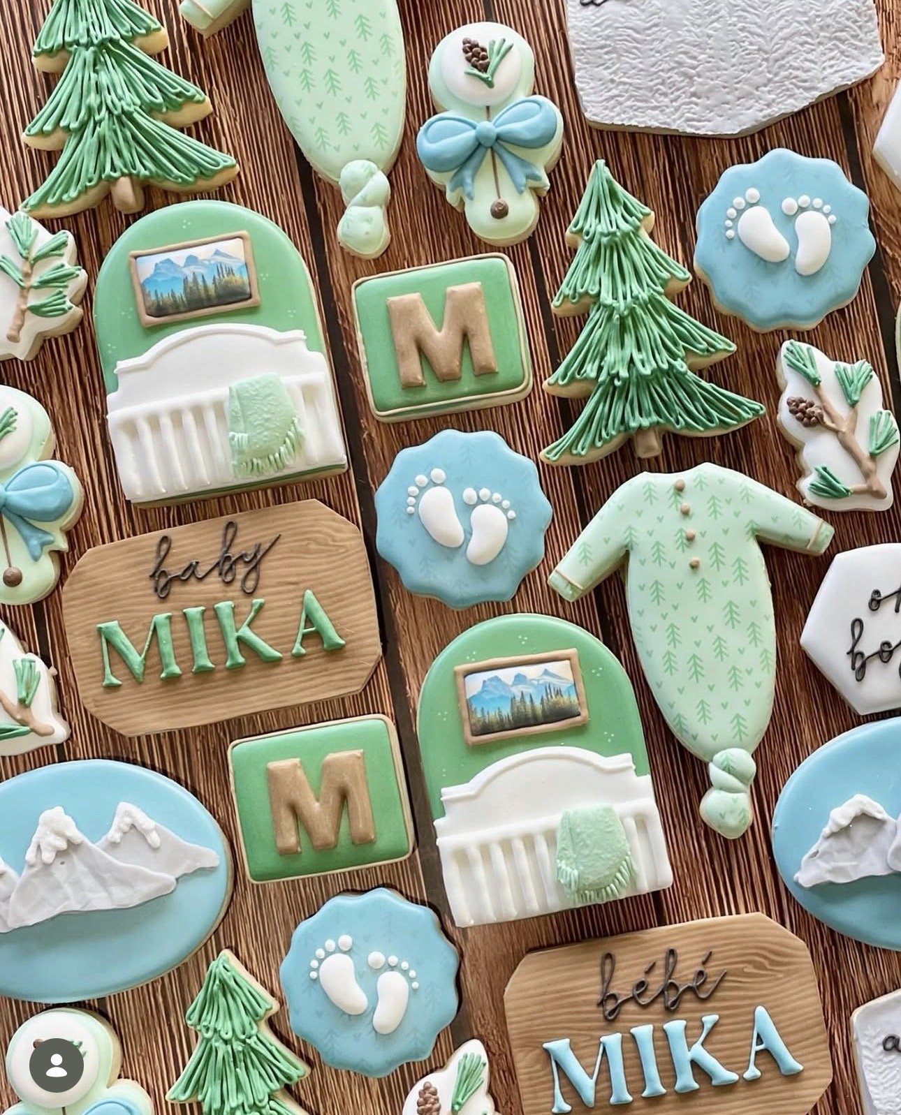 Decorated baby shower cookies featuring pine trees, mountains, baby footprints, sailboats, onesies, rattles, and personalized “Mika” plaques in blue, green, and white.