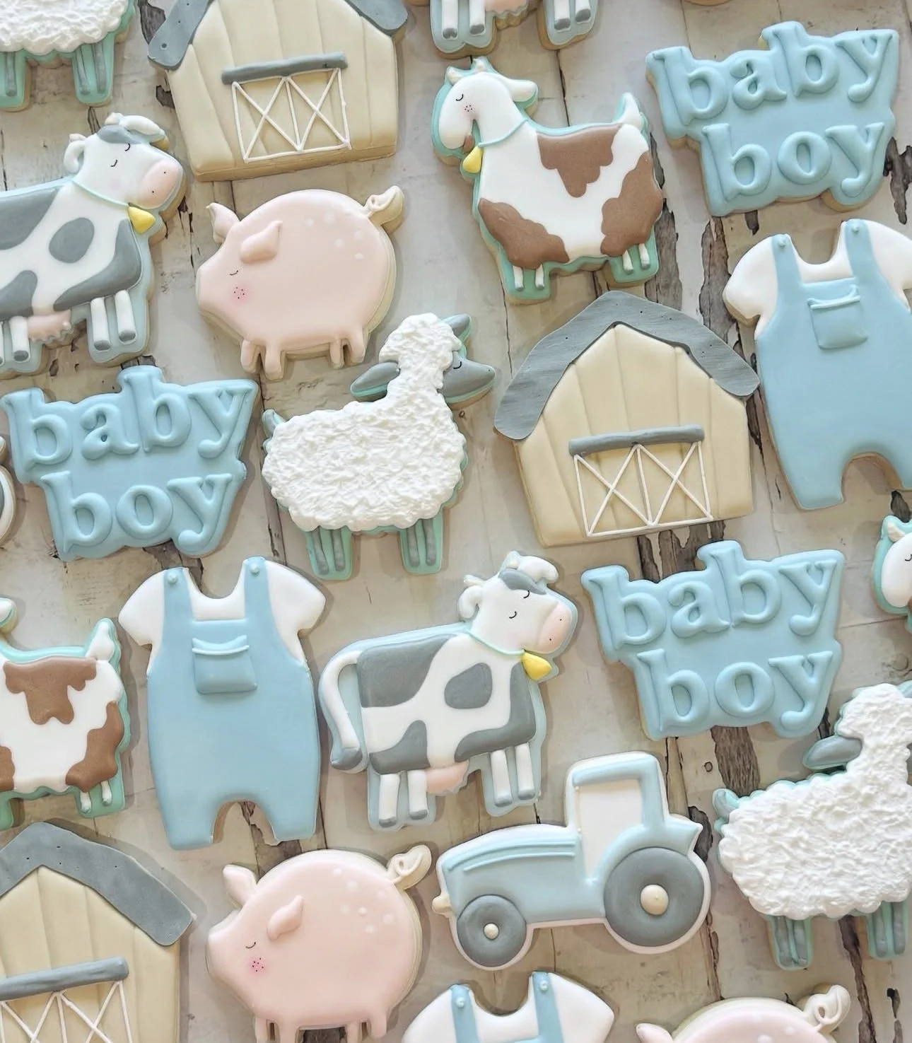 Pastel farm-themed baby shower cookies featuring cows, pigs, sheep, tractors, barns, overalls, and blue “baby boy” lettering