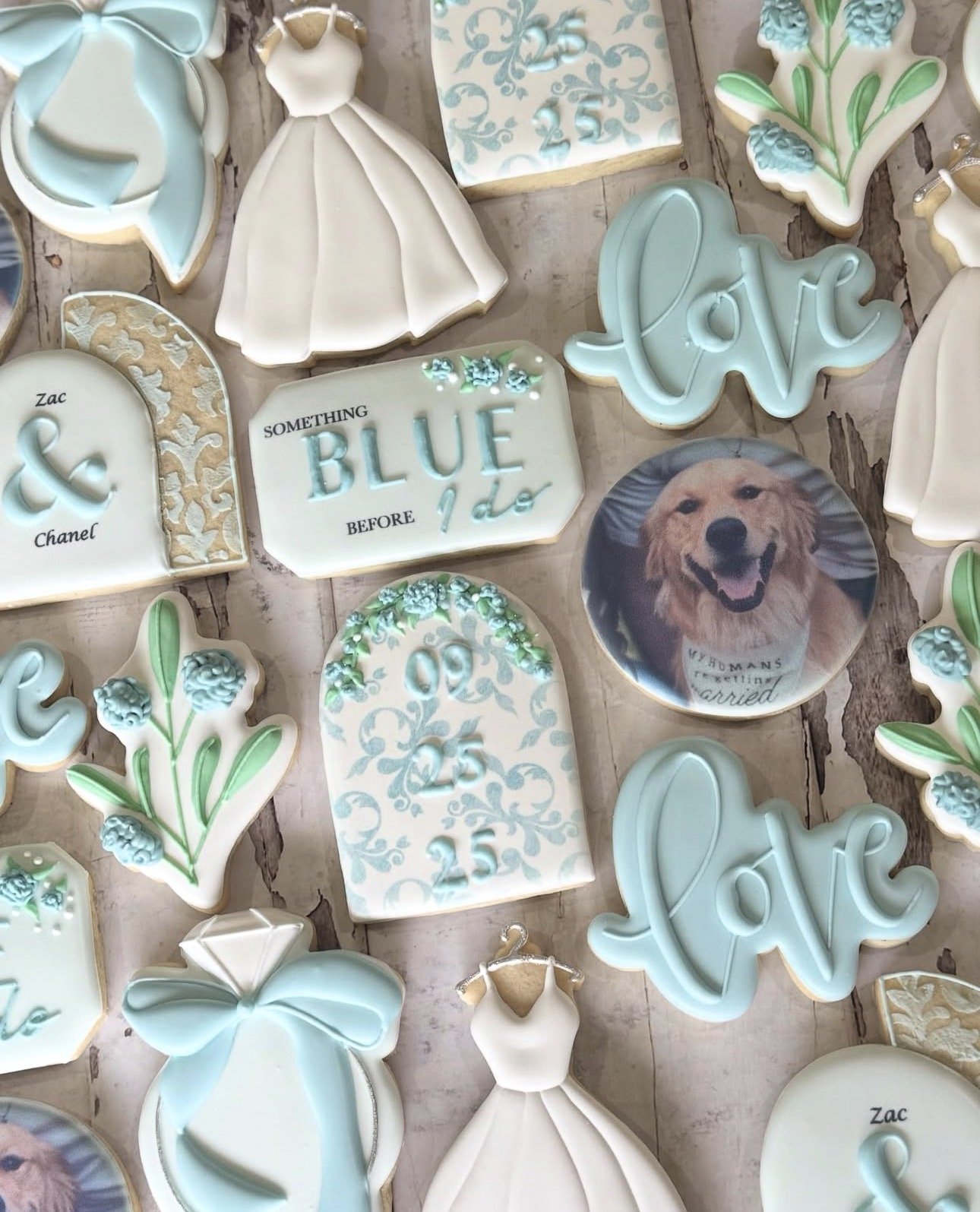 Assortment of pastel blue and white decorated cookies, including wedding dresses, floral sprigs, names, monograms, and a golden retriever portrait cookie.