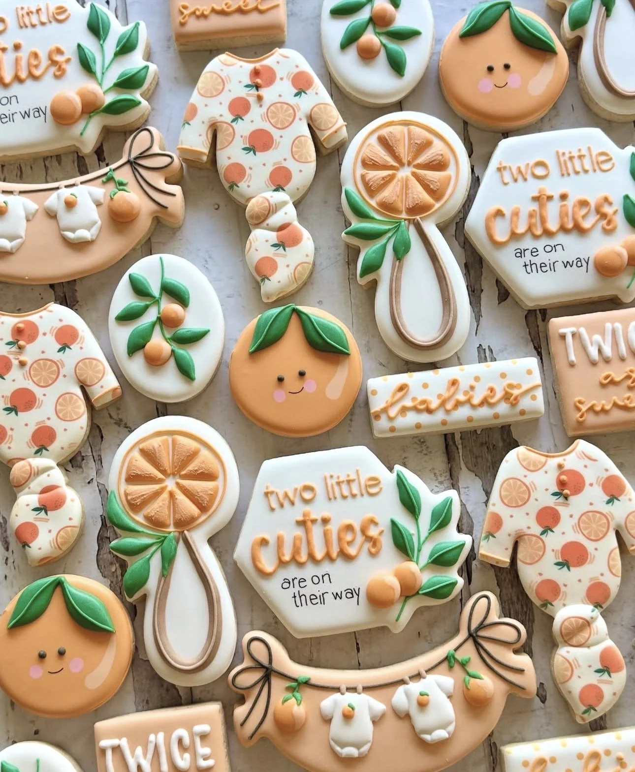 Decorated citrus-themed baby shower cookies arranged on a rustic wood surface, including oranges, citrus slices, baby onesies, and cookies reading “Two little cuties are on their way.”