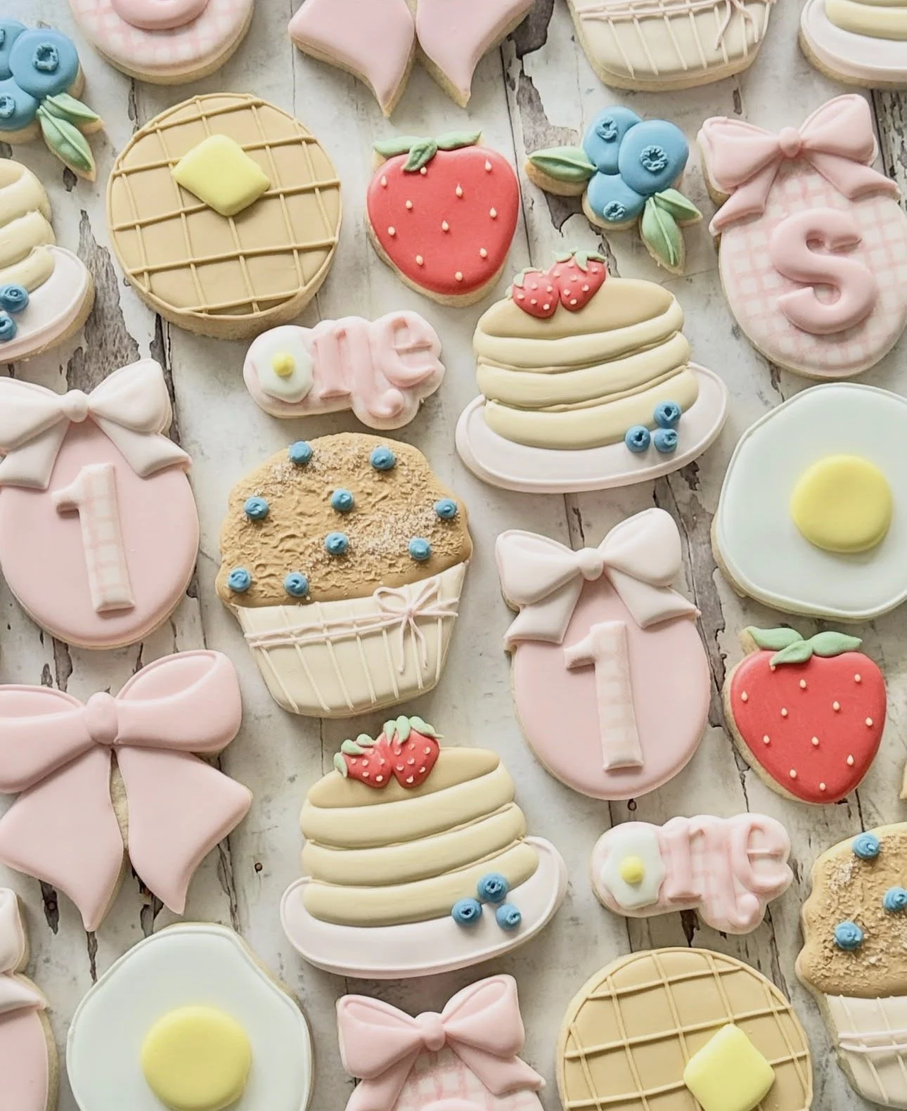 Pastel decorated sugar cookies arranged on a rustic wooden surface, including strawberries, bows, cakes, blueberries, eggs, and baby-themed designs.