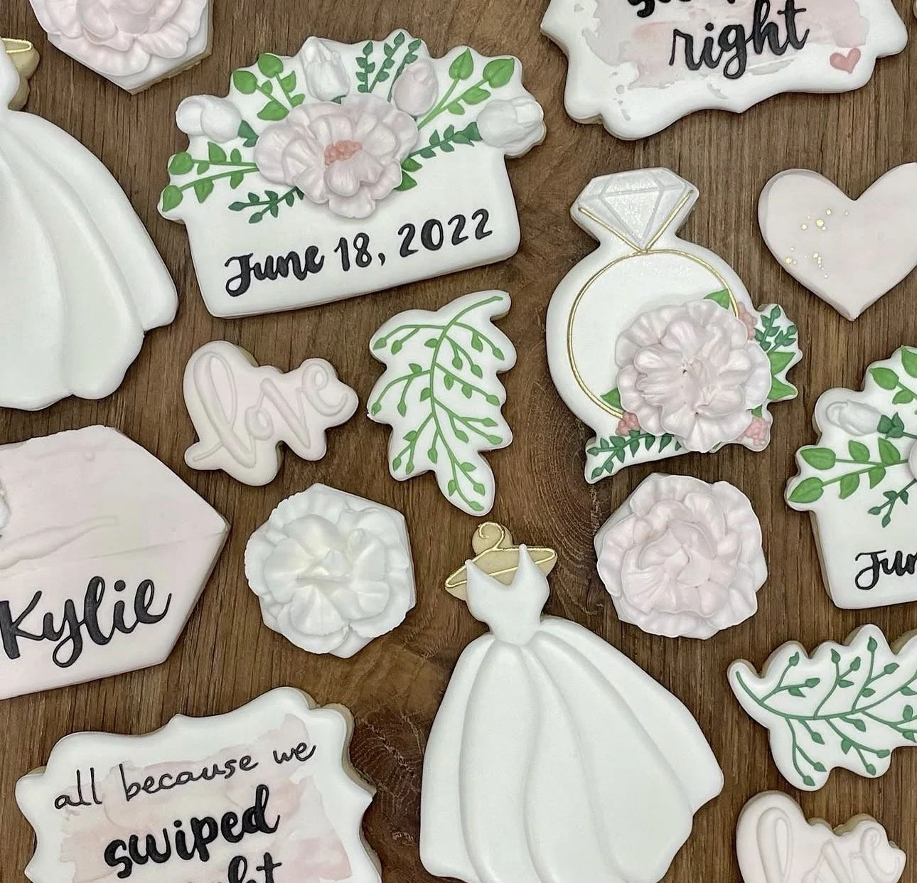Decorated wedding cookies on a wooden surface, including floral designs, a diamond ring, wedding dress, greenery, hearts, and personalized messages.