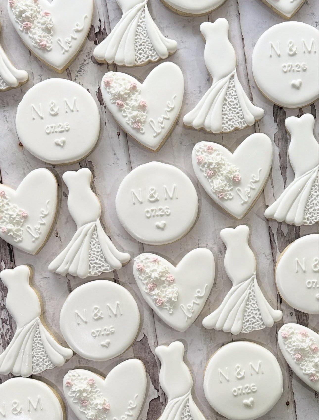 White wedding-themed sugar cookies on a rustic wood background, including heart-shaped floral cookies, wedding dress cookies, and round cookies reading “N&M 07.26.”