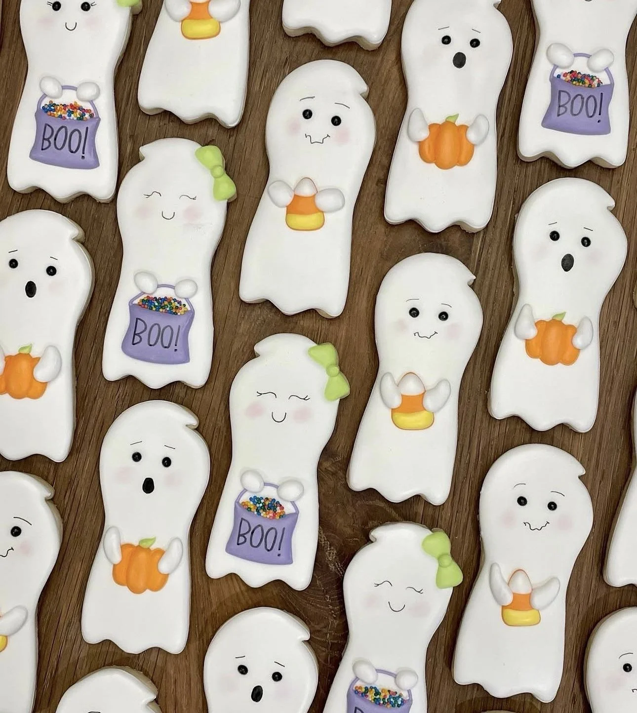 Halloween ghost cookies decorated with pumpkins, candy corn, green bows, and purple BOO candy bags on a wooden surface