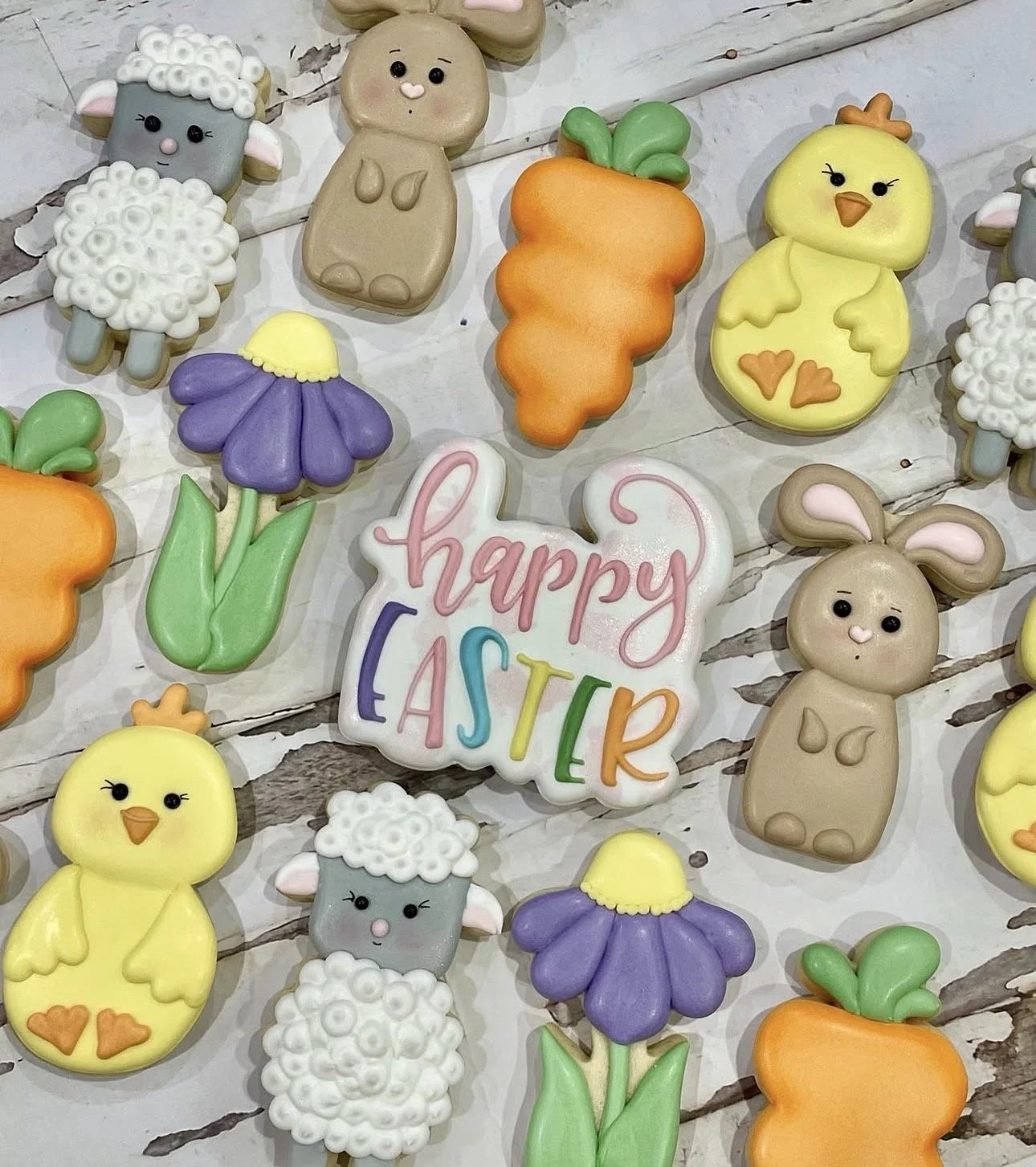 Colorful Easter-themed cookies shaped like chicks, lambs, bunnies, carrots, flowers, and a “Happy Easter” sign on a rustic white wooden background.