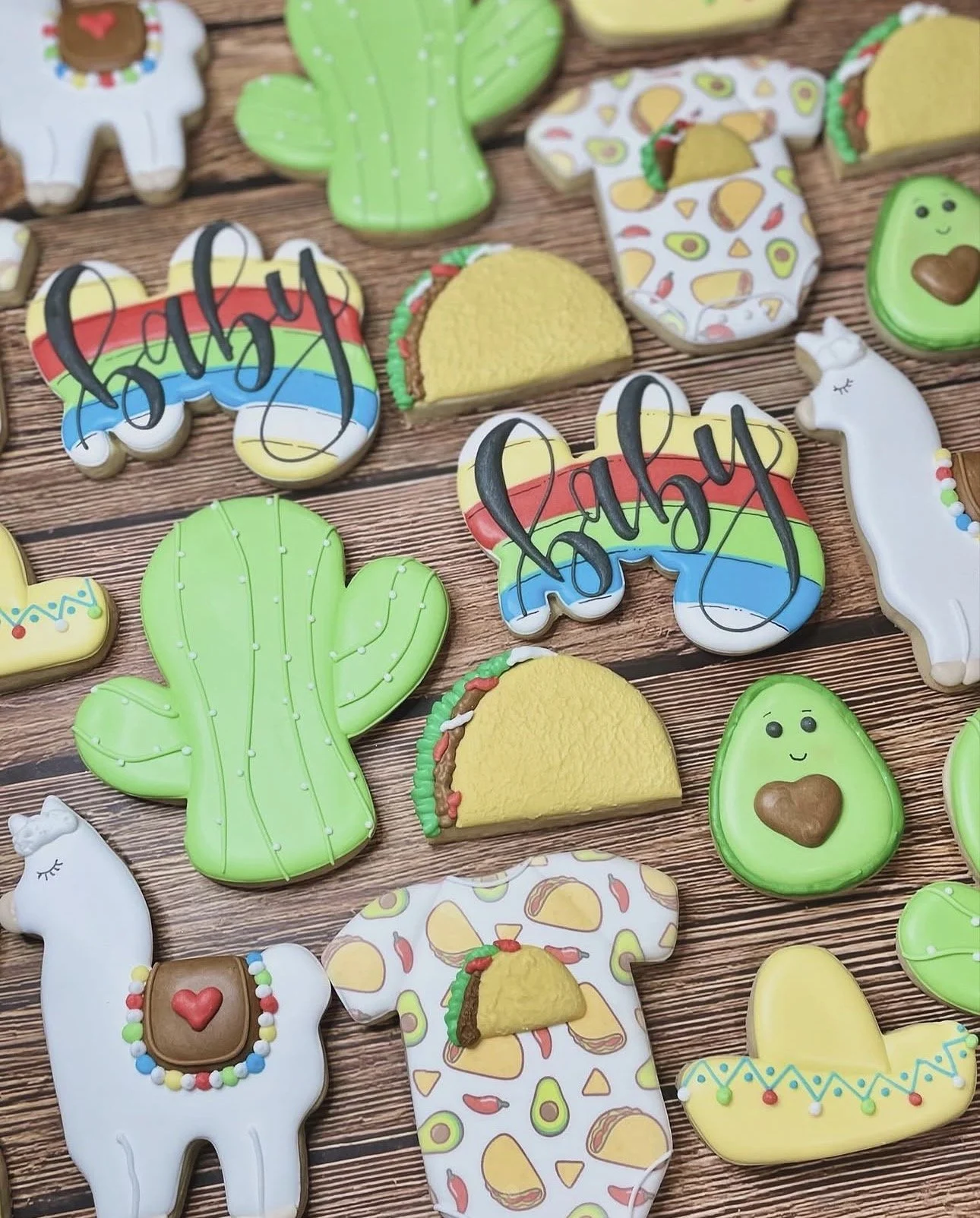 Colorful taco-themed decorated cookies featuring cacti, avocados, llamas, sombreros, and rainbow designs on a wooden background