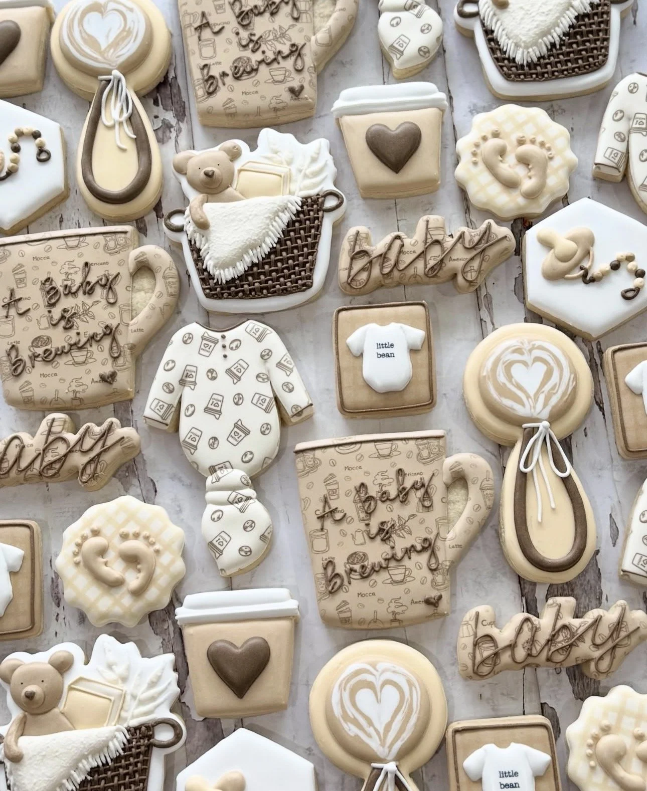 Assorted coffee- and baby-themed decorated sugar cookies arranged on a rustic wood background, including latte art, mugs, baby clothes, teddy bears, and hearts.
