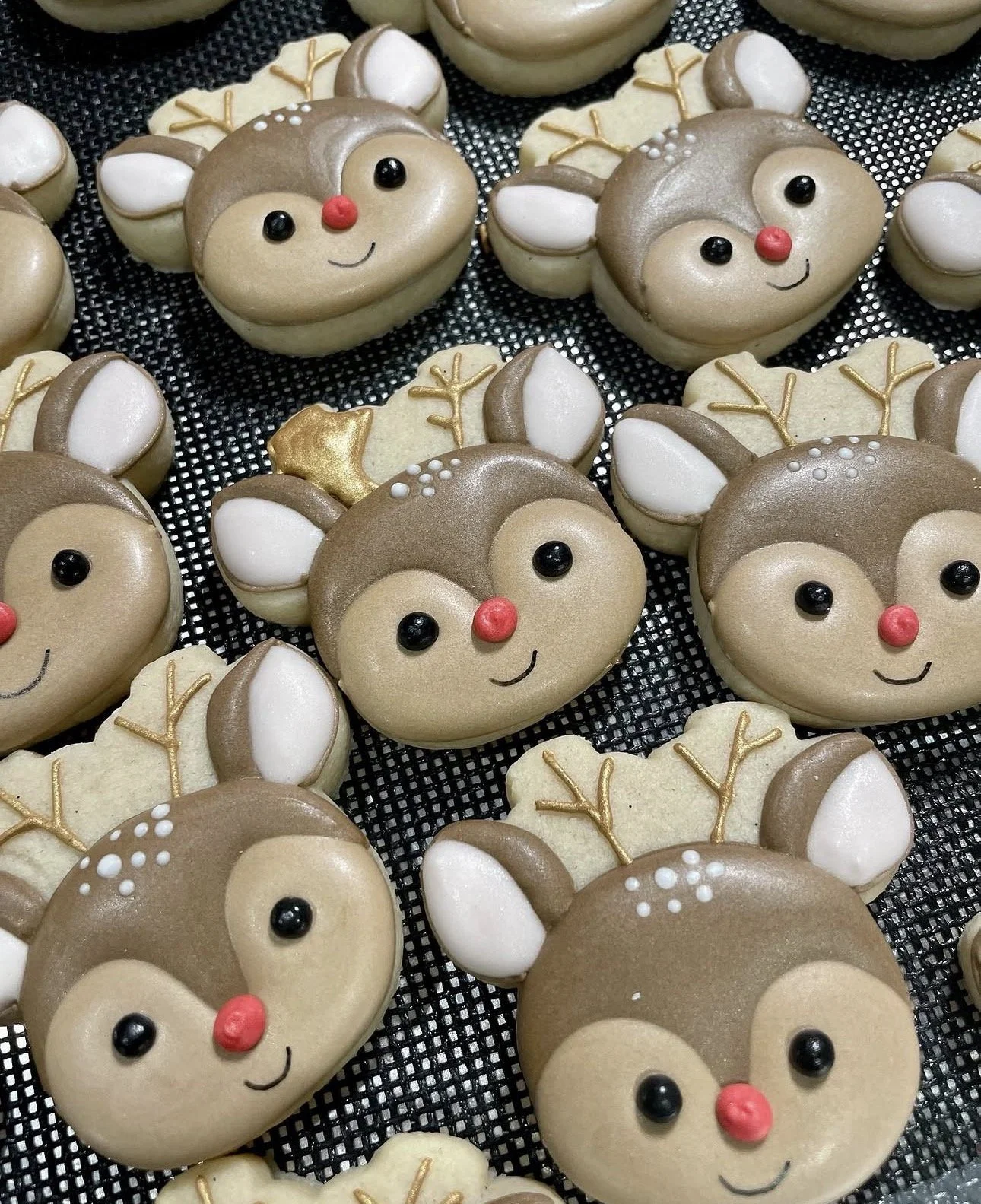 Decorated reindeer macarons with red noses, black eyes, white ears, and antlers arranged on a black baking mat.