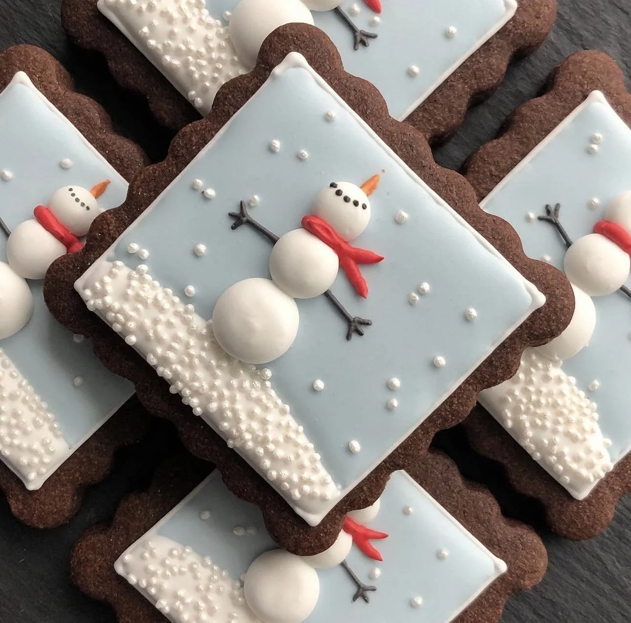 Decorated chocolate snowman cookies with light blue icing, white sprinkles, red scarves, and orange carrot noses