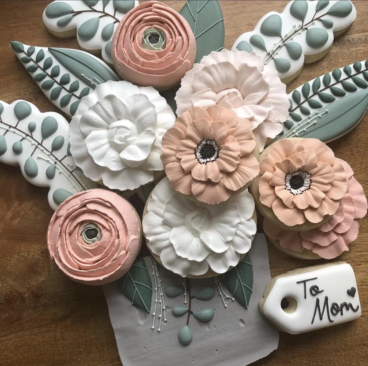 Decorated floral cookies in pink, white, and sage green, arranged with leaves and a “To Mom” gift tag.