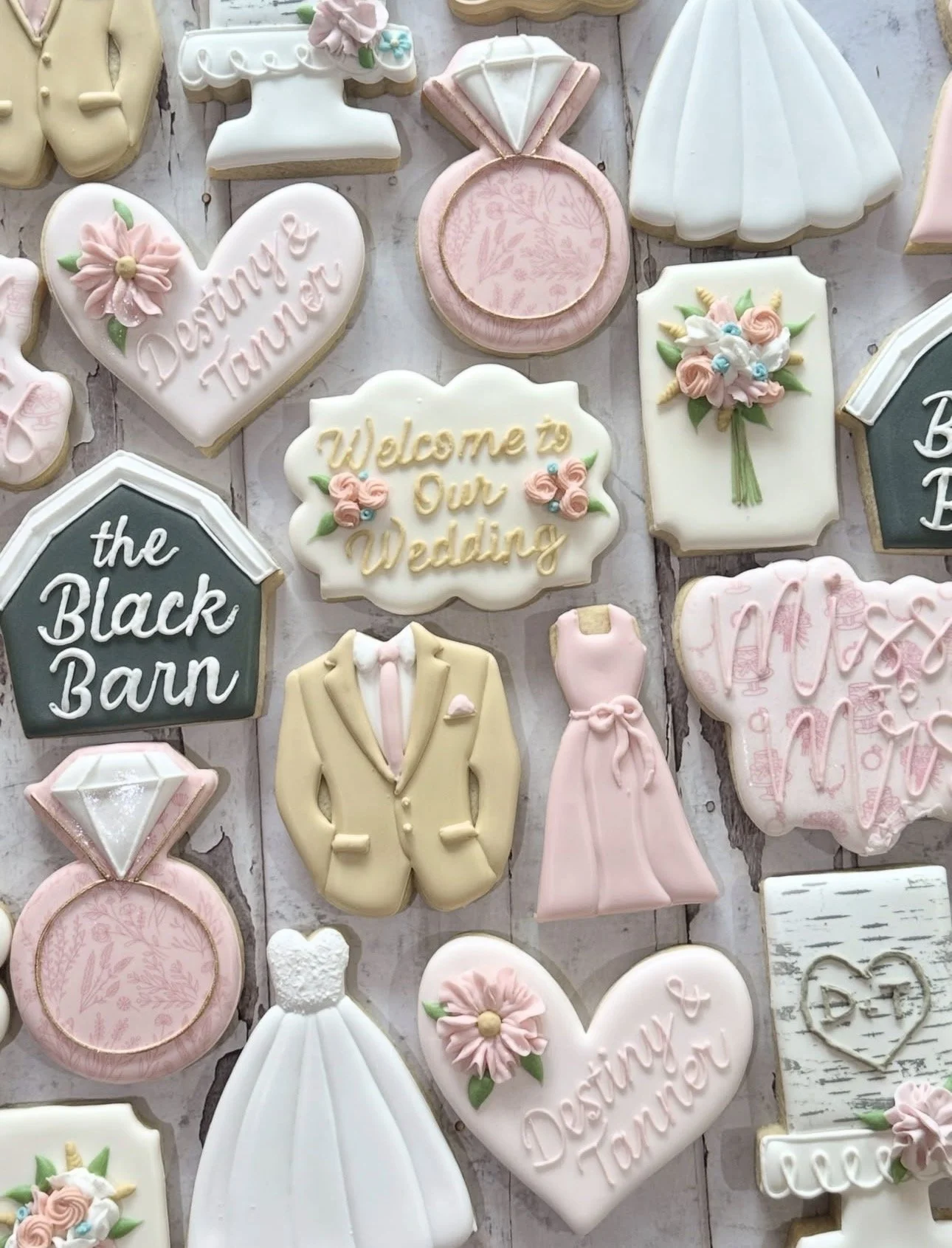 Decorated wedding-themed sugar cookies featuring dresses, suits, diamond rings, bouquets, and messages including “Welcome to Our Wedding” and “the Black Barn.”