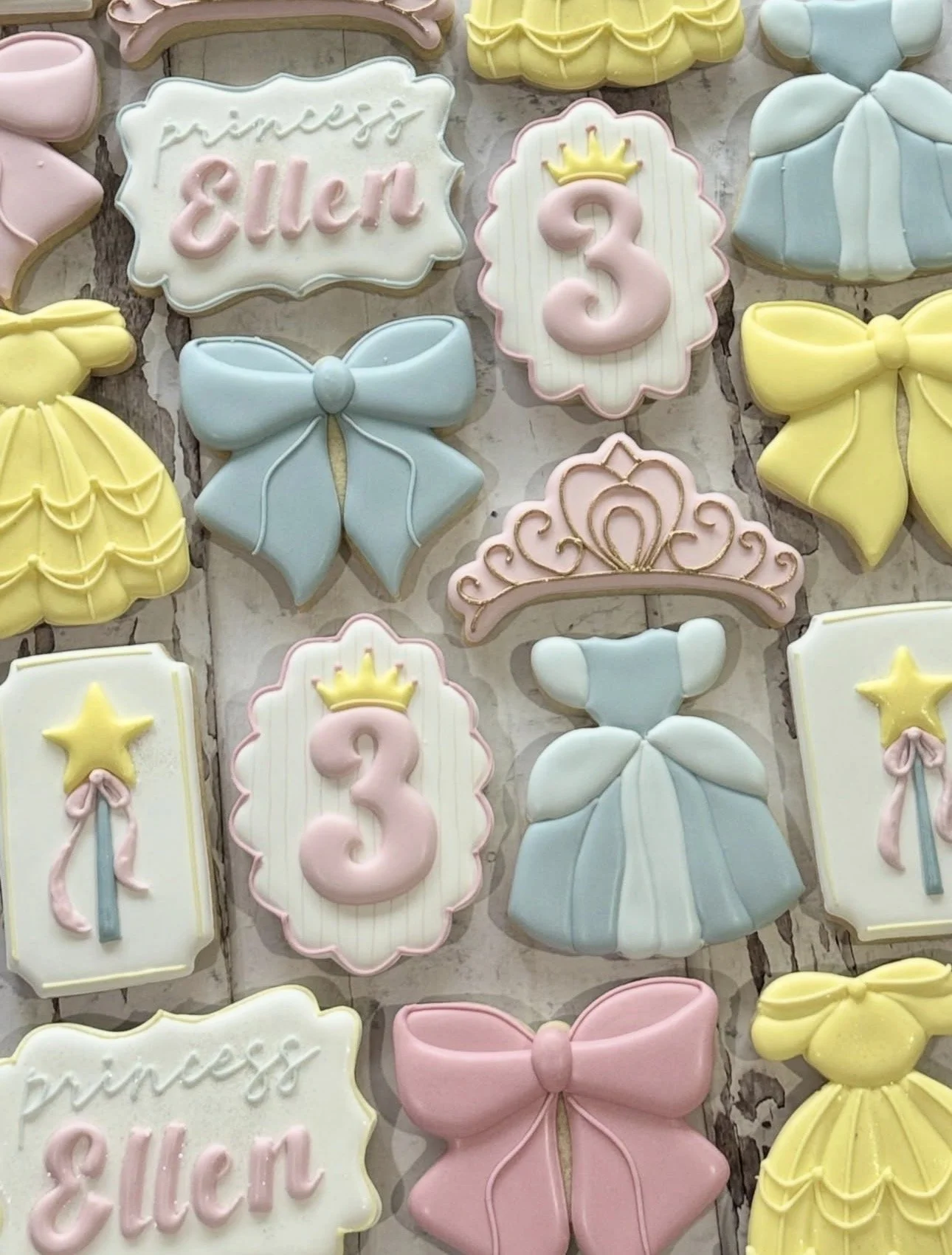 Pastel princess-themed decorated cookies featuring crowns, bows, dresses, magic wands, tiaras, and cookies reading “Princess Eleni” with the number three.