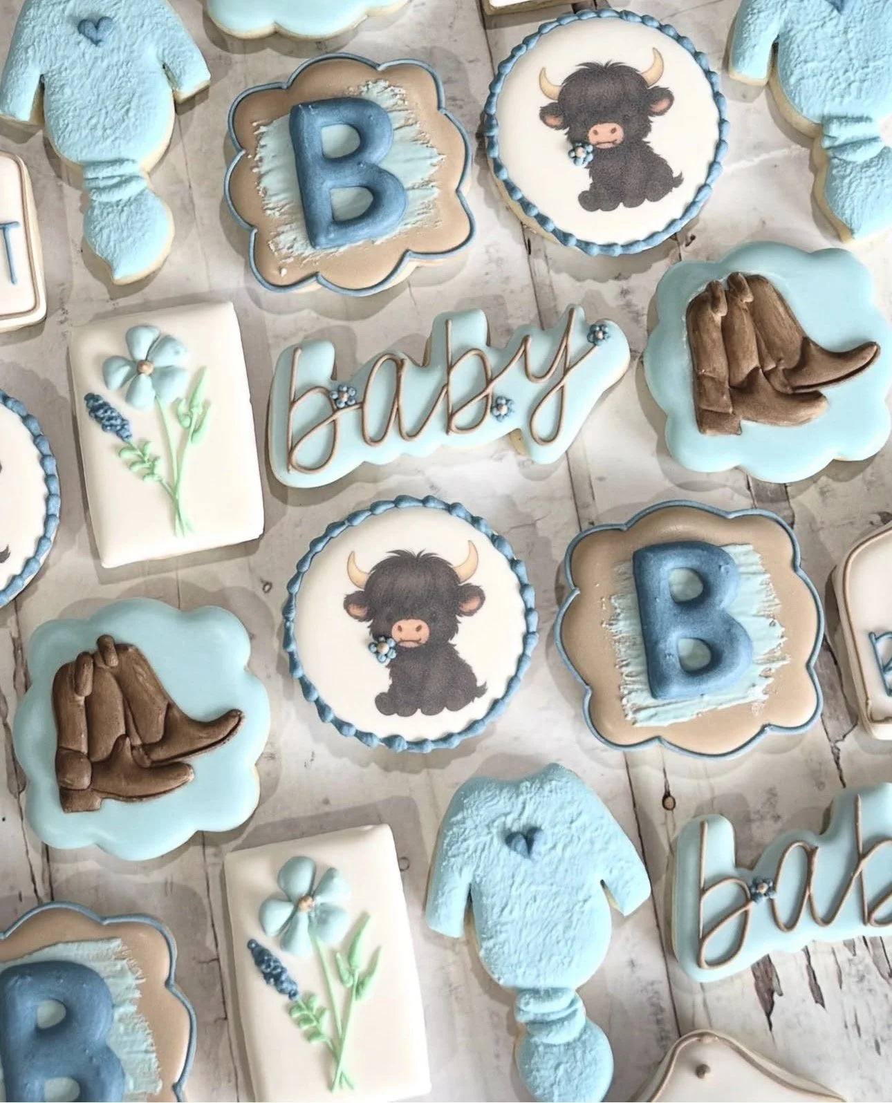 Rustic western-themed decorated sugar cookies featuring Highland cows, cowboy hats, blue baby bodysuits, flowers, and the name “baby”.
