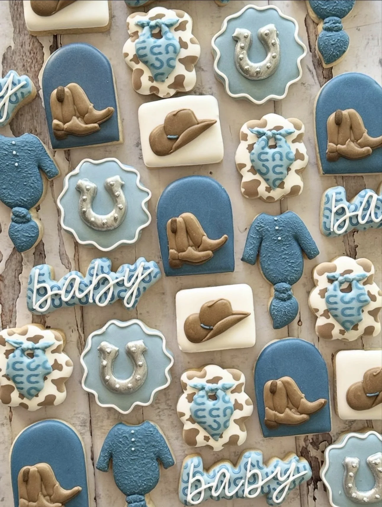 Cowboy-themed decorated cookies with cowboy hats, blue boots, horseshoes, blue shirts, and Western lettering on a rustic wood background