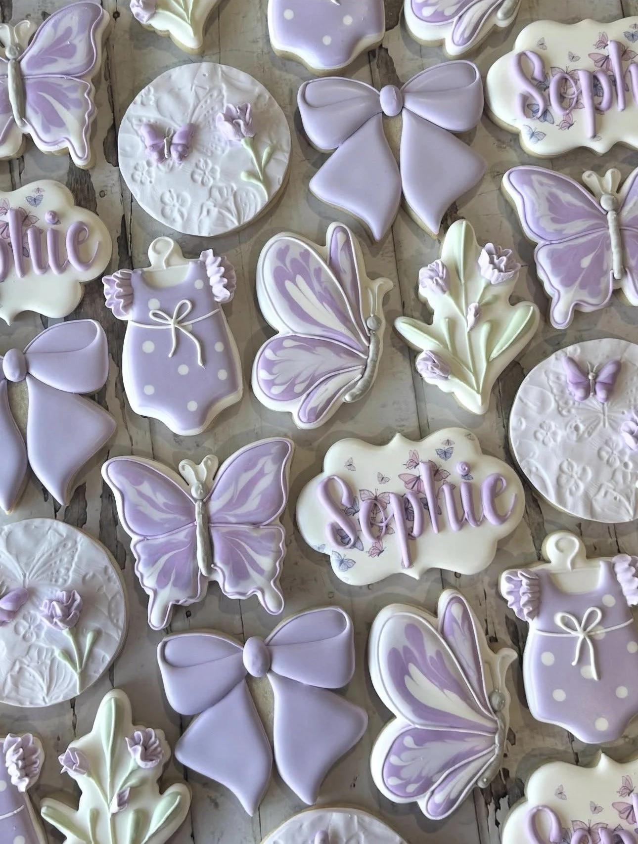 Assortment of pastel purple and white decorated cookies shaped like butterflies, bows, flowers, dresses, and name plaques on a rustic wooden surface