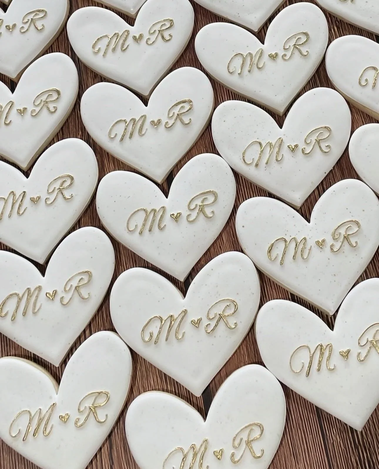 White heart-shaped cookies decorated with gold initials “M ♥ R” on a wood-grain surface