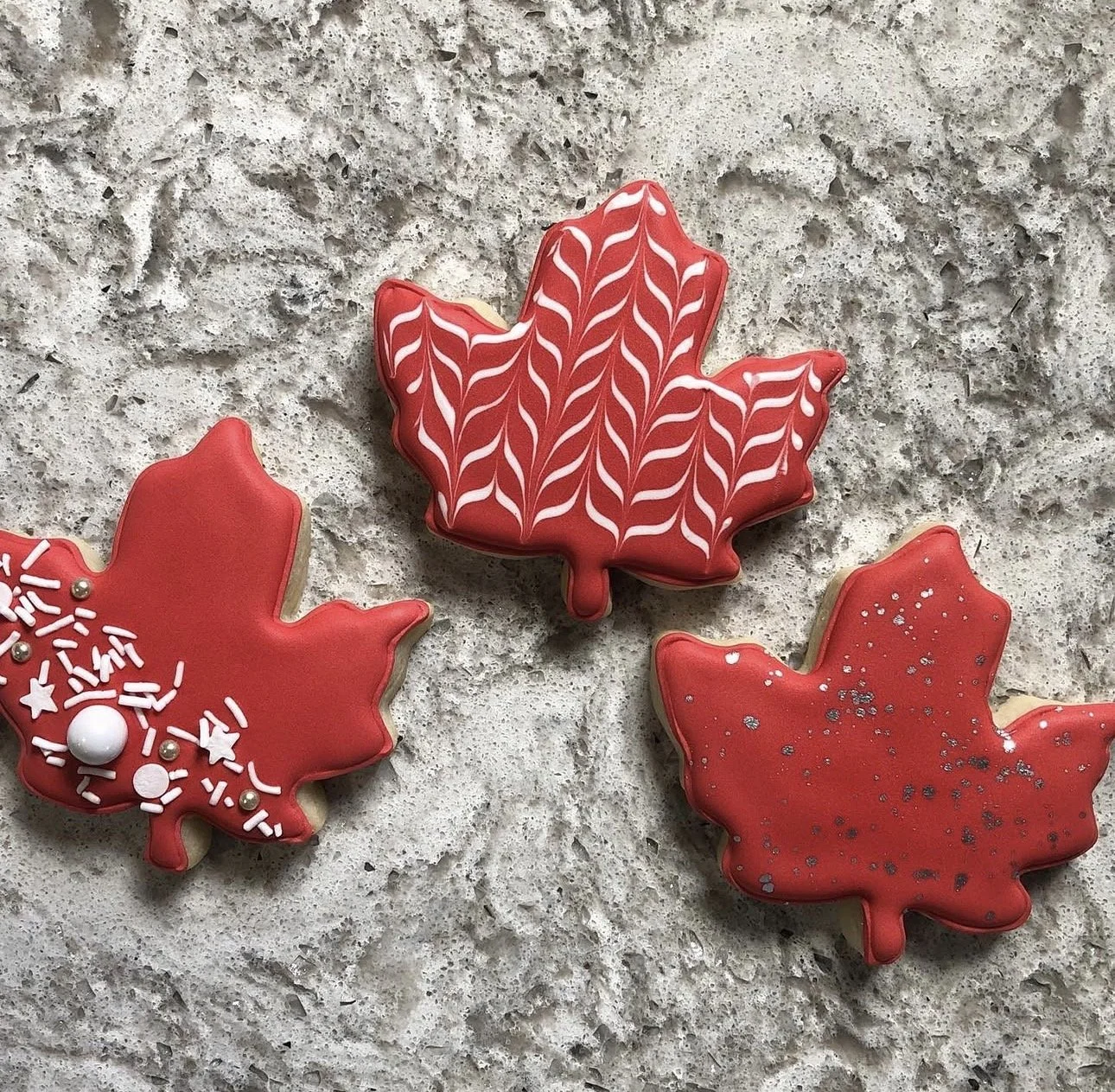 Three red maple leaf-shaped cookies with white icing, sprinkles, and glitter on a gray stone surface
