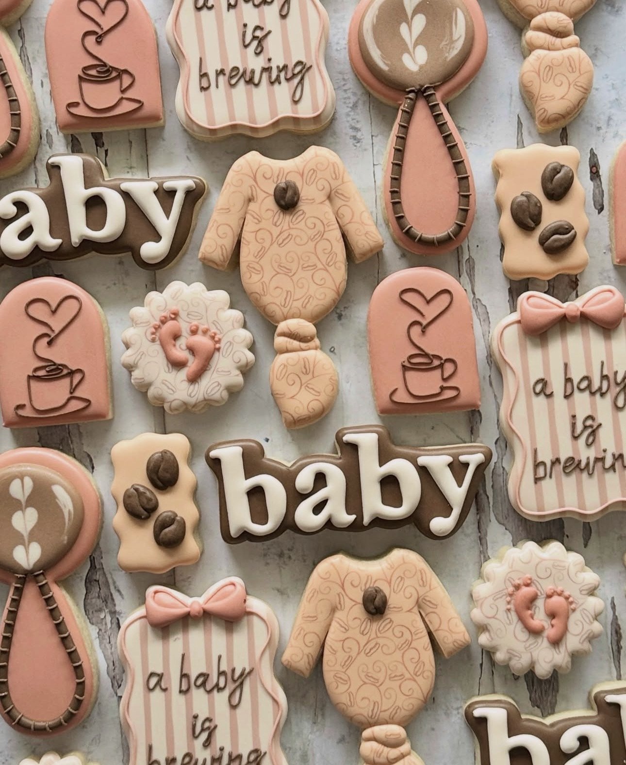 Decorated baby shower cookies featuring coffee cups, baby onesies, pacifiers, baby feet, coffee beans, hearts, and messages reading “a baby is brewing.”