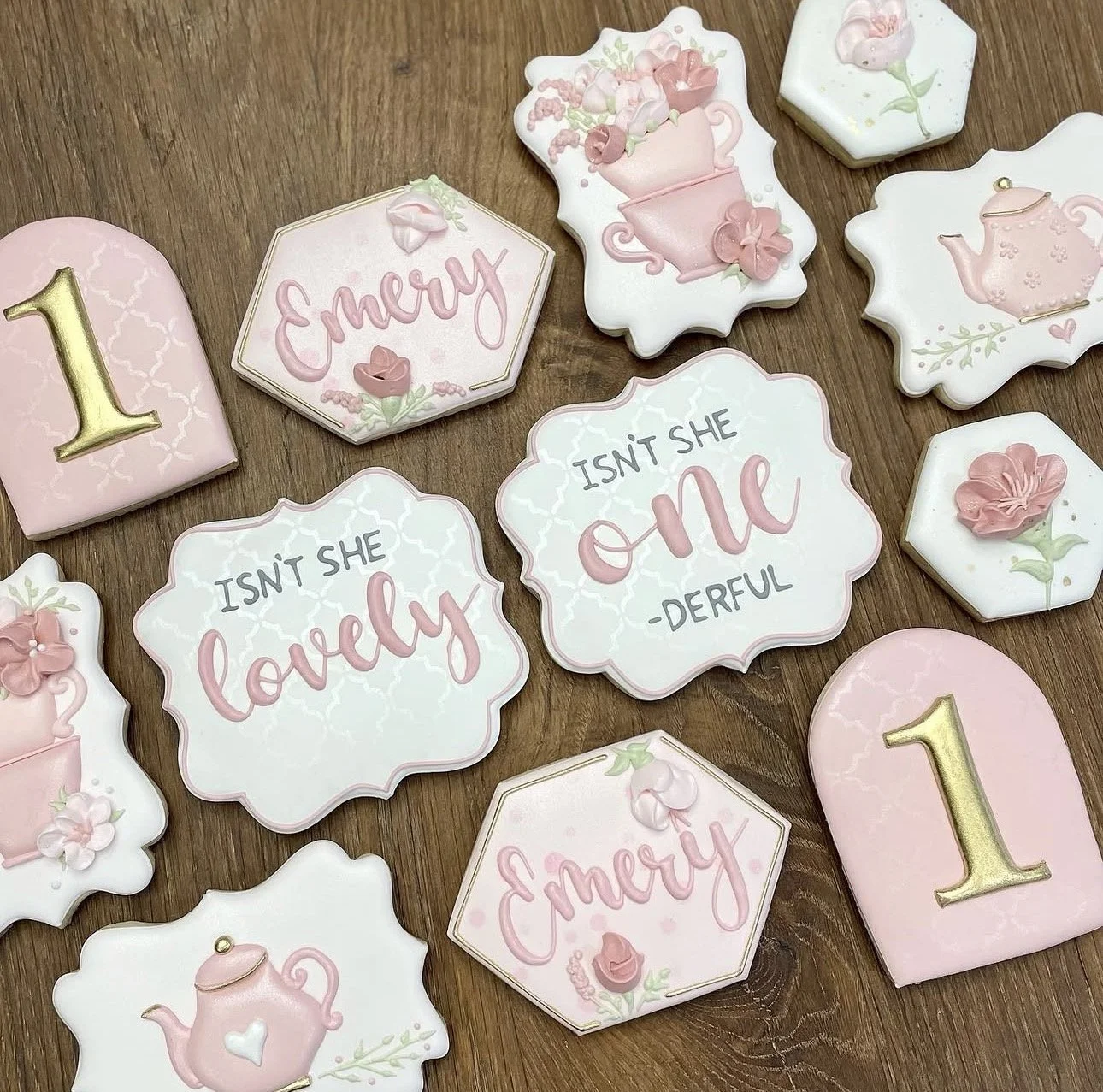 Decorated pink and white baby shower cookies featuring teapots, flowers, the number one, and “Emery” name plaques