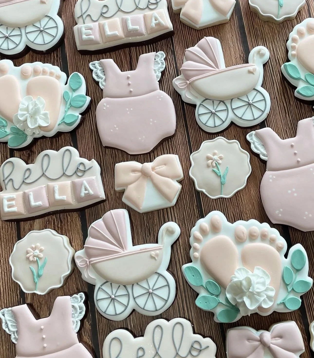 Pastel pink and white baby shower cookies arranged on a wood-grain surface, featuring strollers, onesies, flowers, bows, baby feet, and personalized “Hello Ella” cookies.