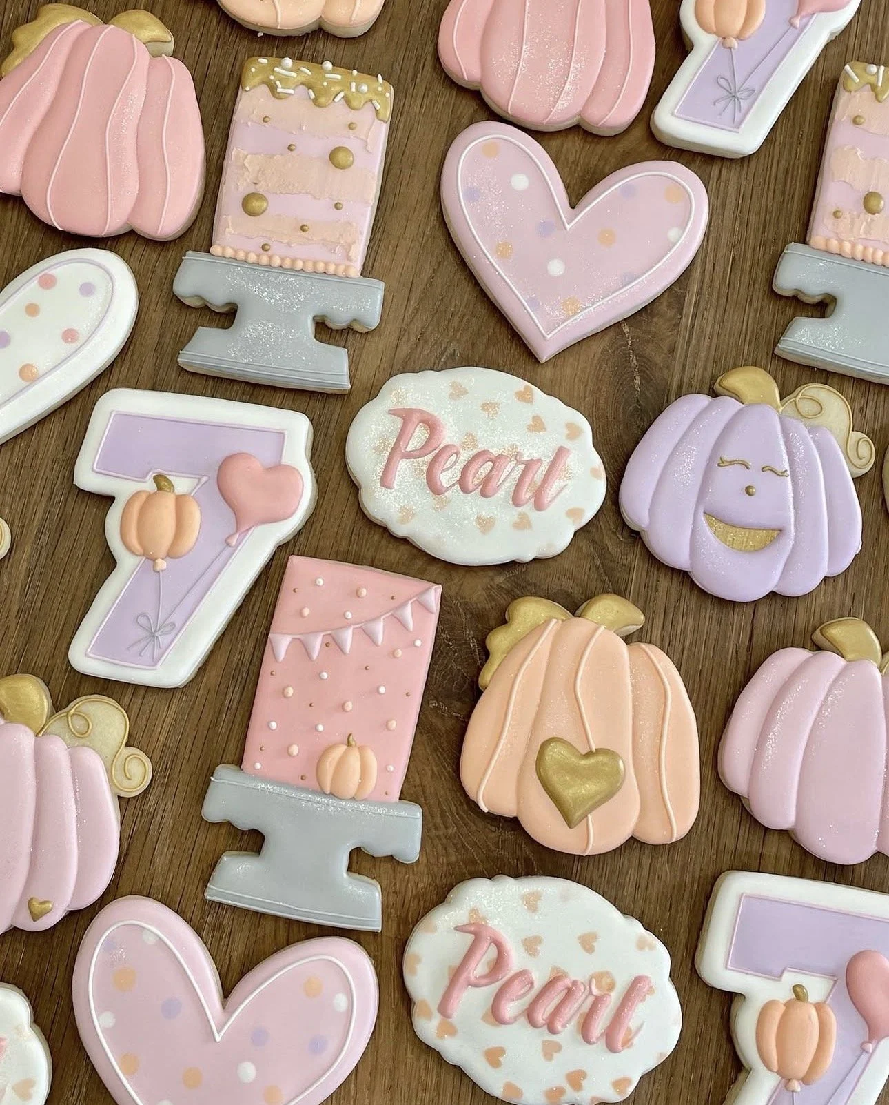 Pastel pumpkin-themed decorated sugar cookies arranged on a wooden table, including hearts, pumpkins, balloons, and cookies spelling “Penny.”