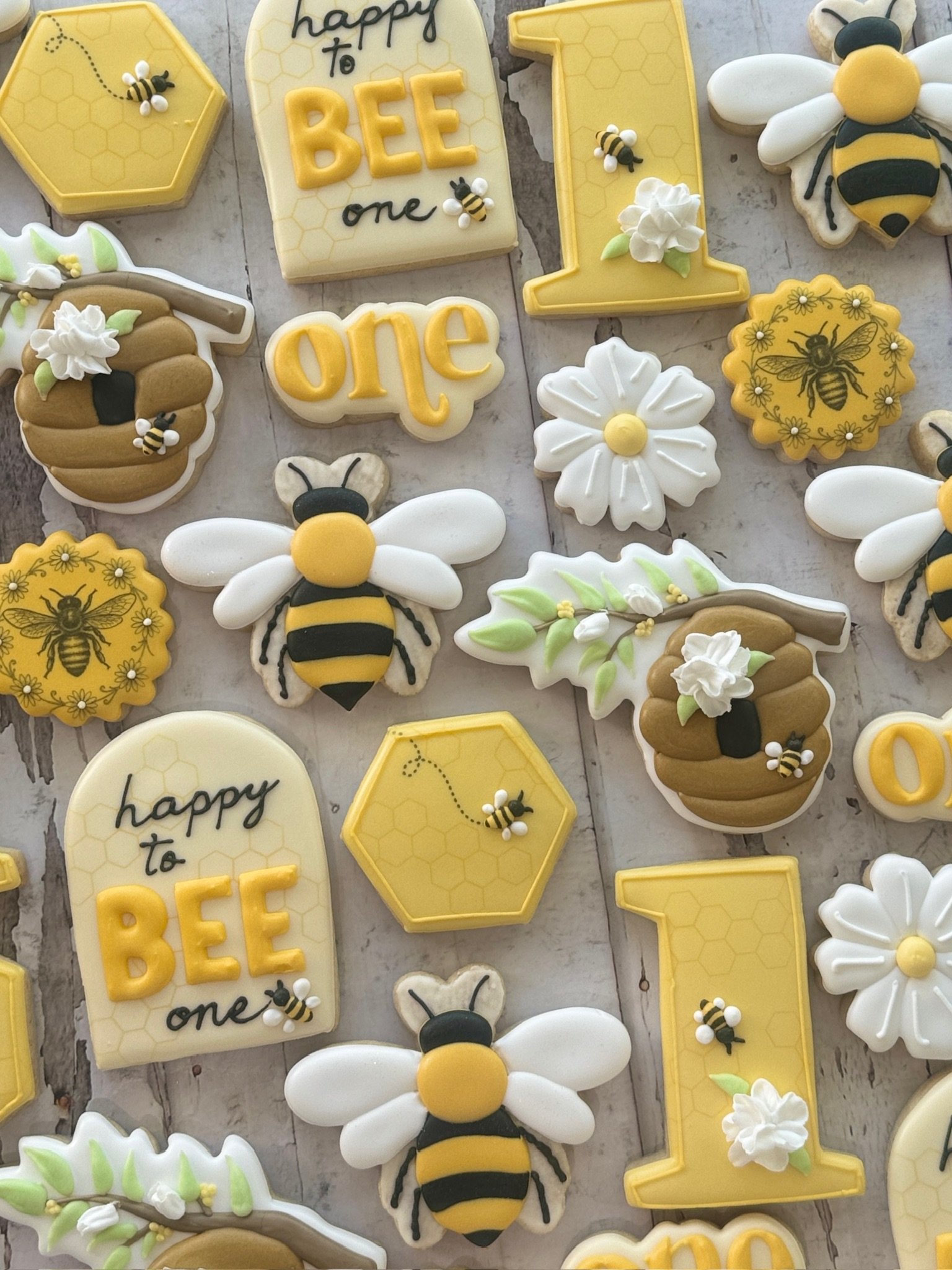 Bee-themed decorated sugar cookies featuring honeycomb hexagons, bees, flowers, beehives, and cookies reading “happy to BEE one.”
