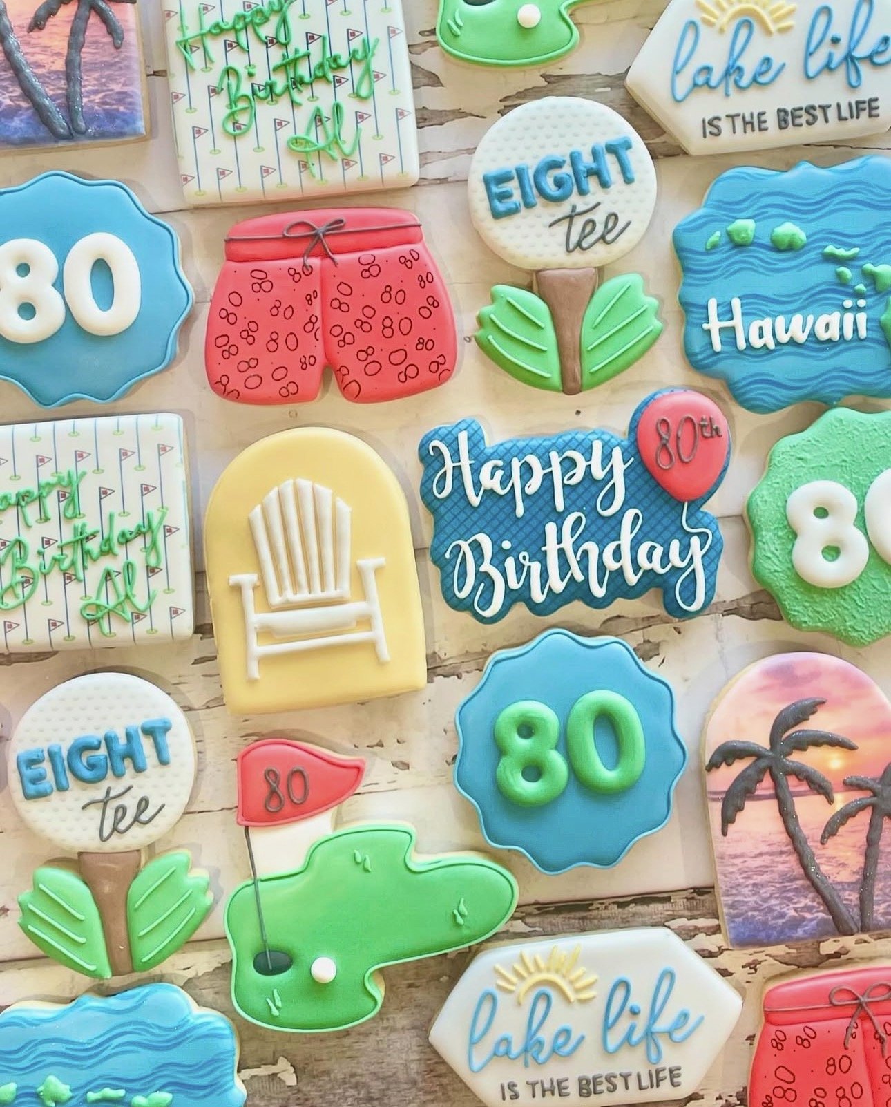 Colorful 80th birthday-themed decorated cookies featuring lake life, Hawaii, golf, palms, Adirondack chairs, alligators, and “Happy Birthday” designs.
