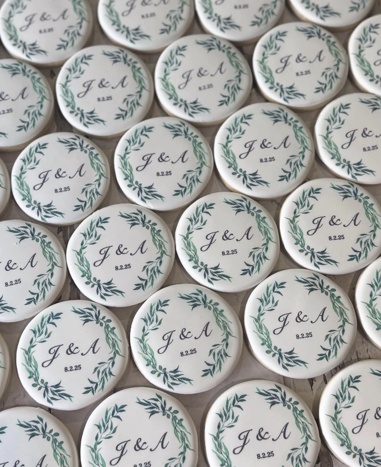 Close-up of personalized wedding cookies decorated with green leafy wreaths, the initials “J & A,” and the date “8.2.25.”