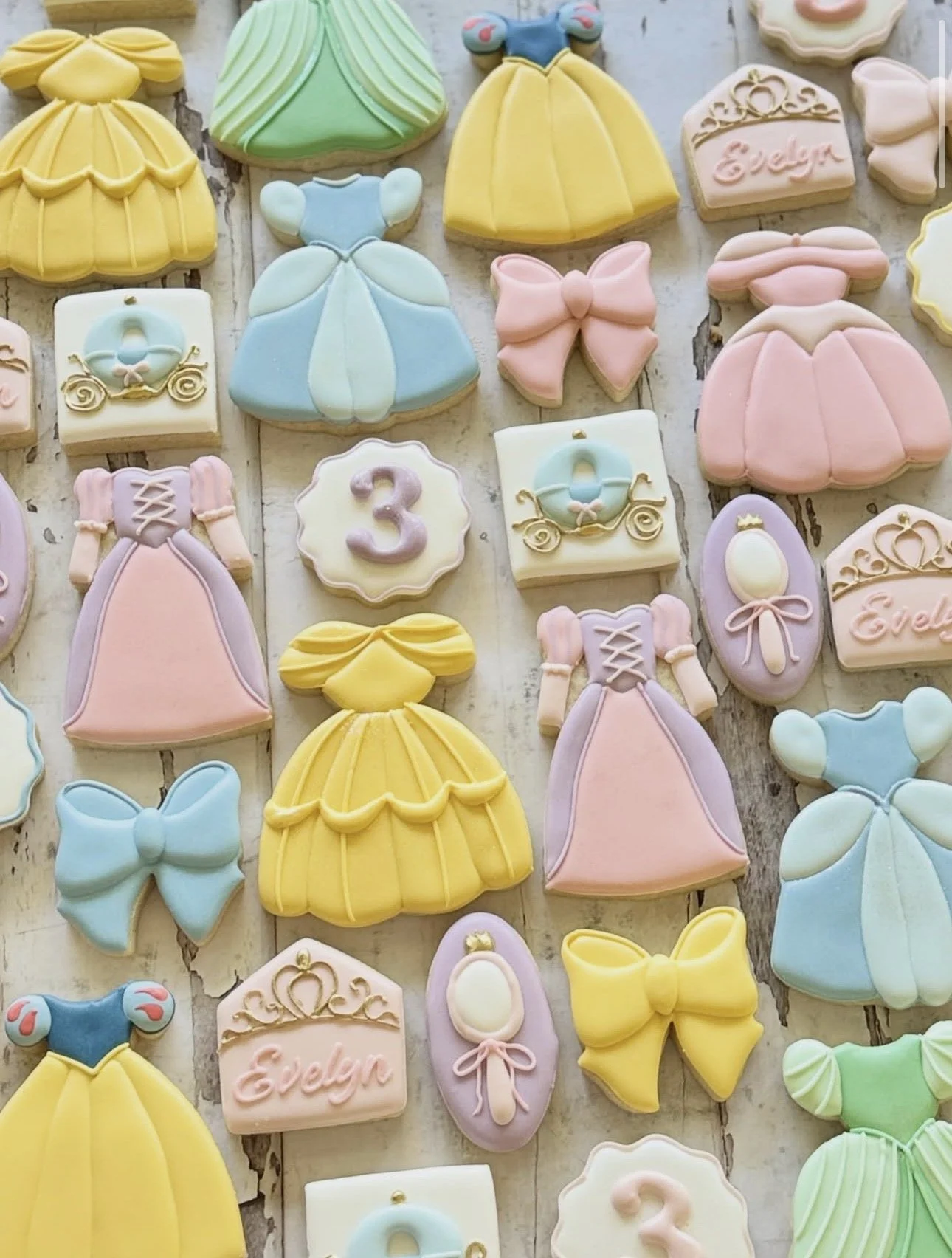 Pastel princess-themed sugar cookies featuring dresses, bows, carriages, crowns, and personalized “Evelyn” cookies arranged on a rustic wooden surface.