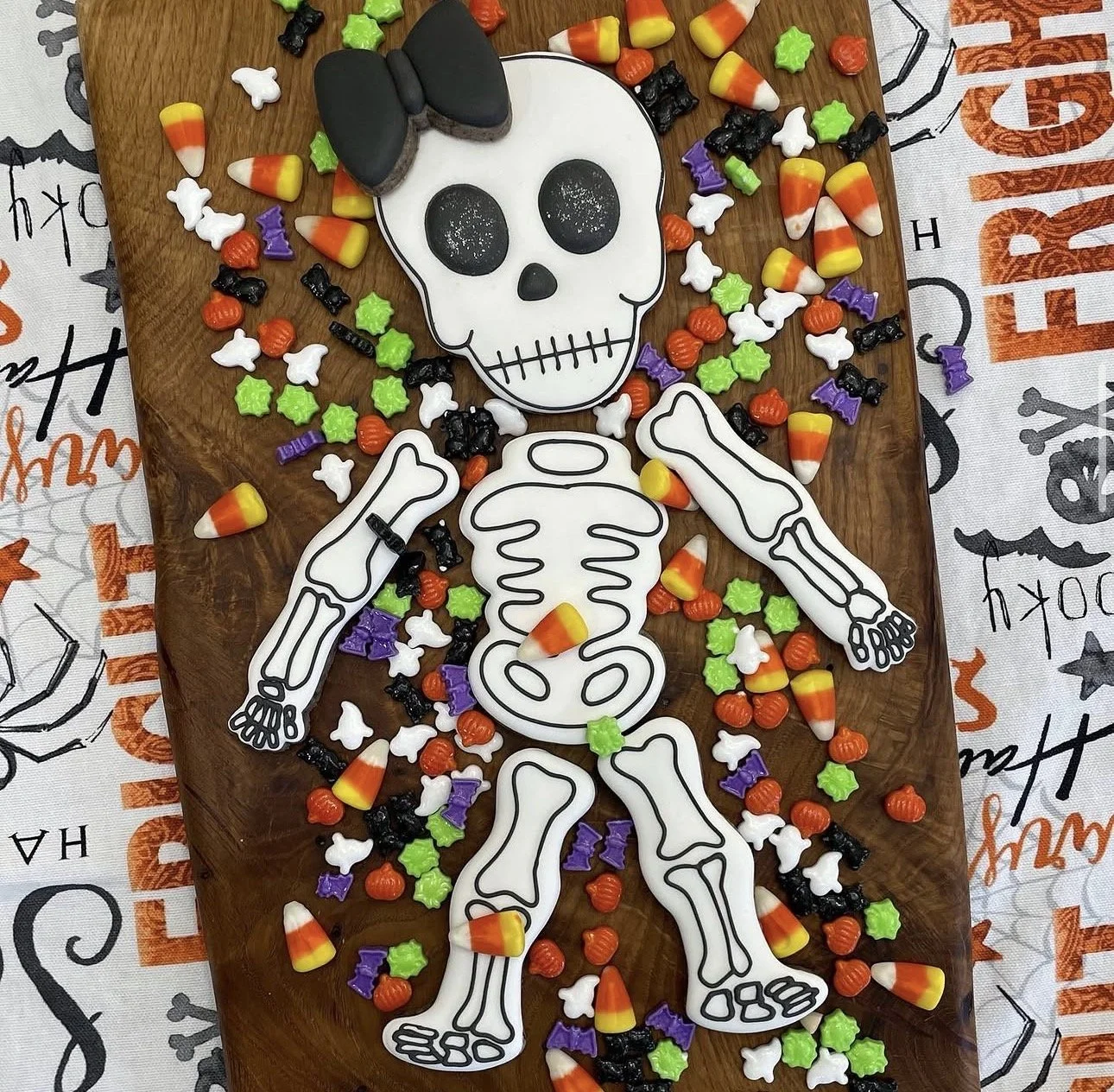 Halloween skeleton cookie topped with a black bow, surrounded by candy corn and colorful Halloween candies on a wooden board