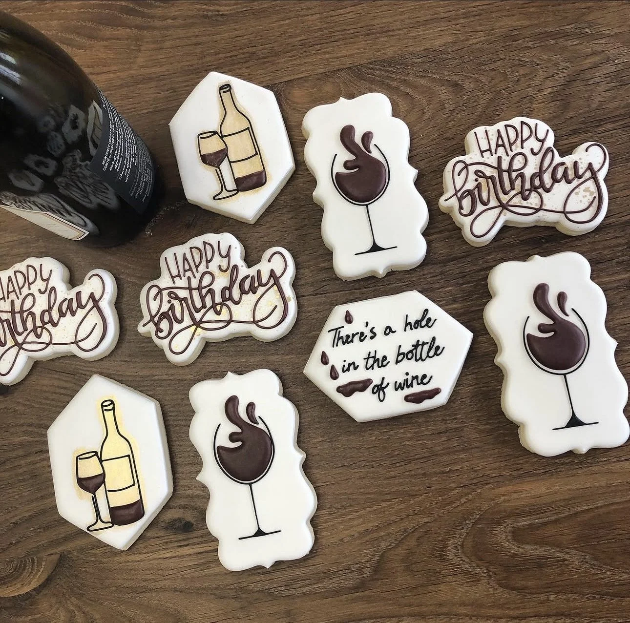 Wine-themed decorated sugar cookies on a wooden table, featuring wine bottles, red wine glasses, and birthday messages.
