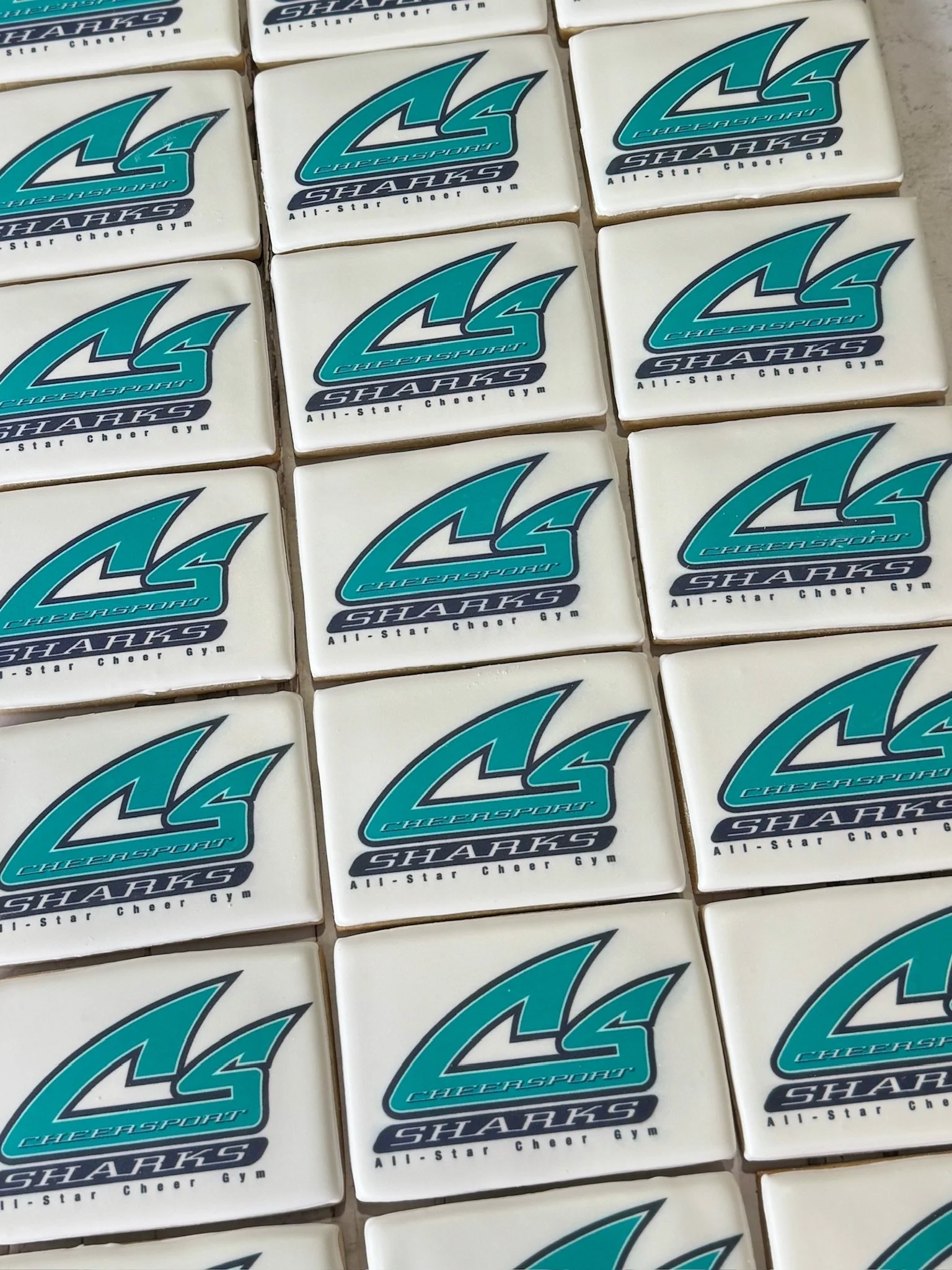 Rows of white cookies decorated with the teal and black Sharks All-Star Cheer Gym logo.