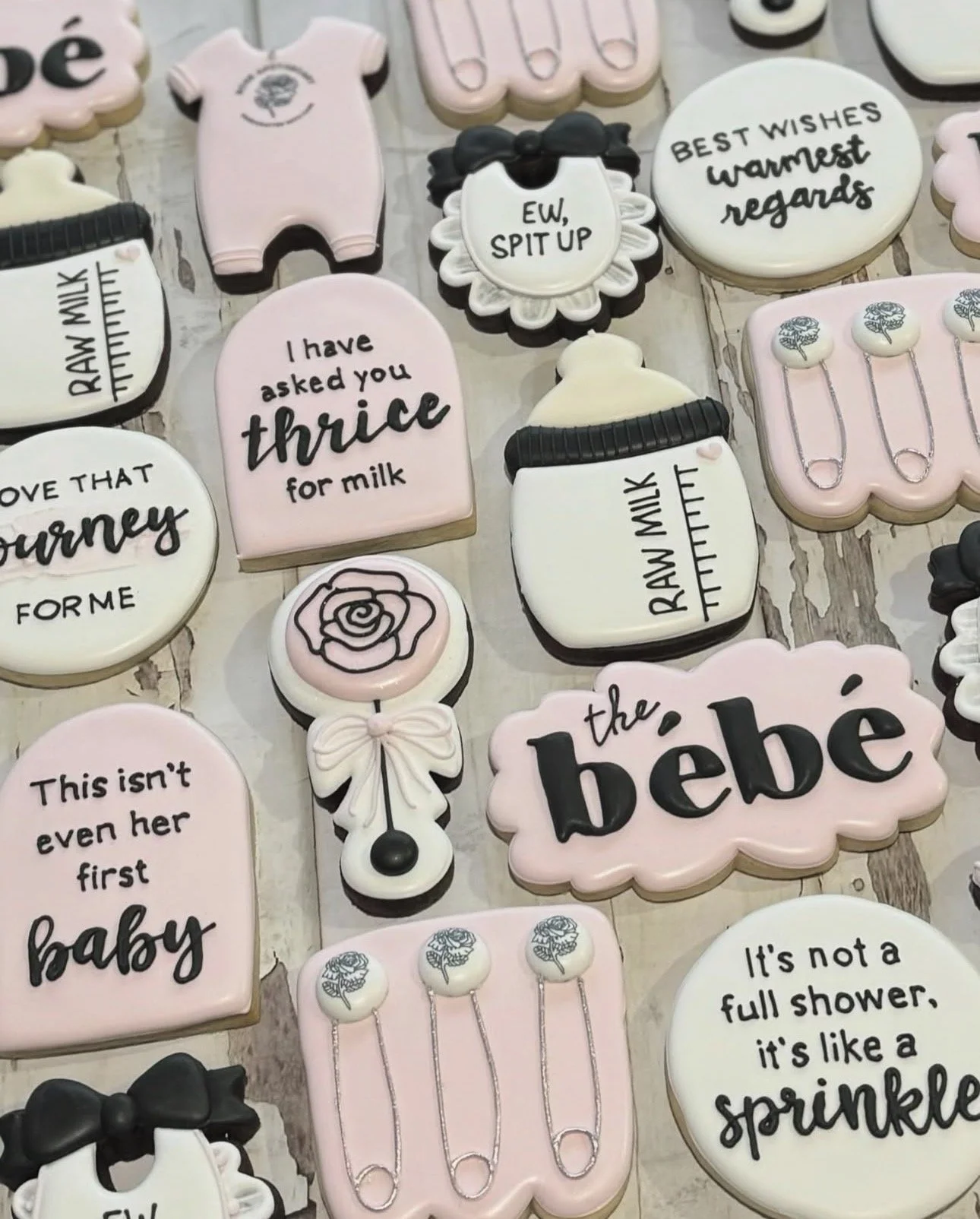 Assorted pink and white baby shower cookies decorated with baby clothes, milk bottles, safety pins, pacifiers, bows, and humorous messages
