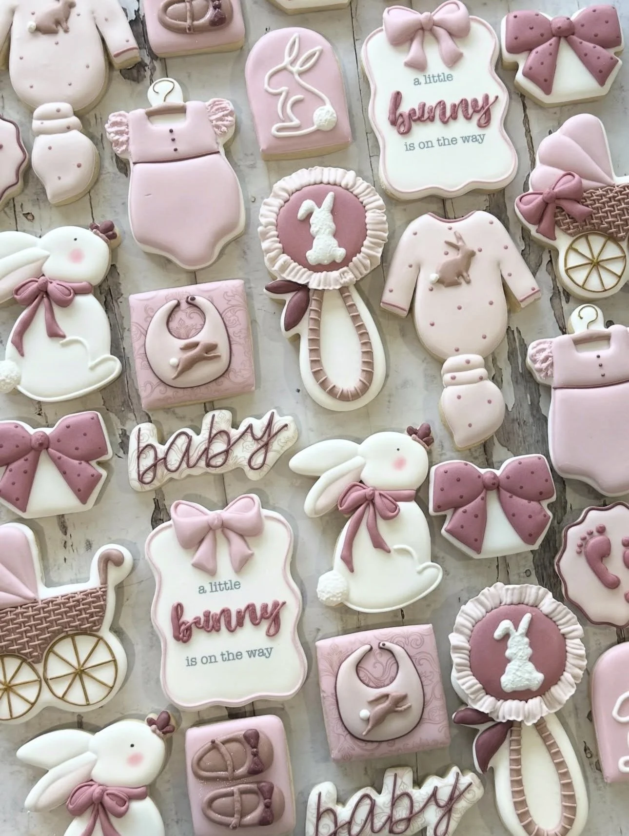 Pastel pink and white baby shower cookies featuring bunnies, onesies, bows, baby bottles, rattles, pacifiers, strollers, and “a little bunny is on the way” text.