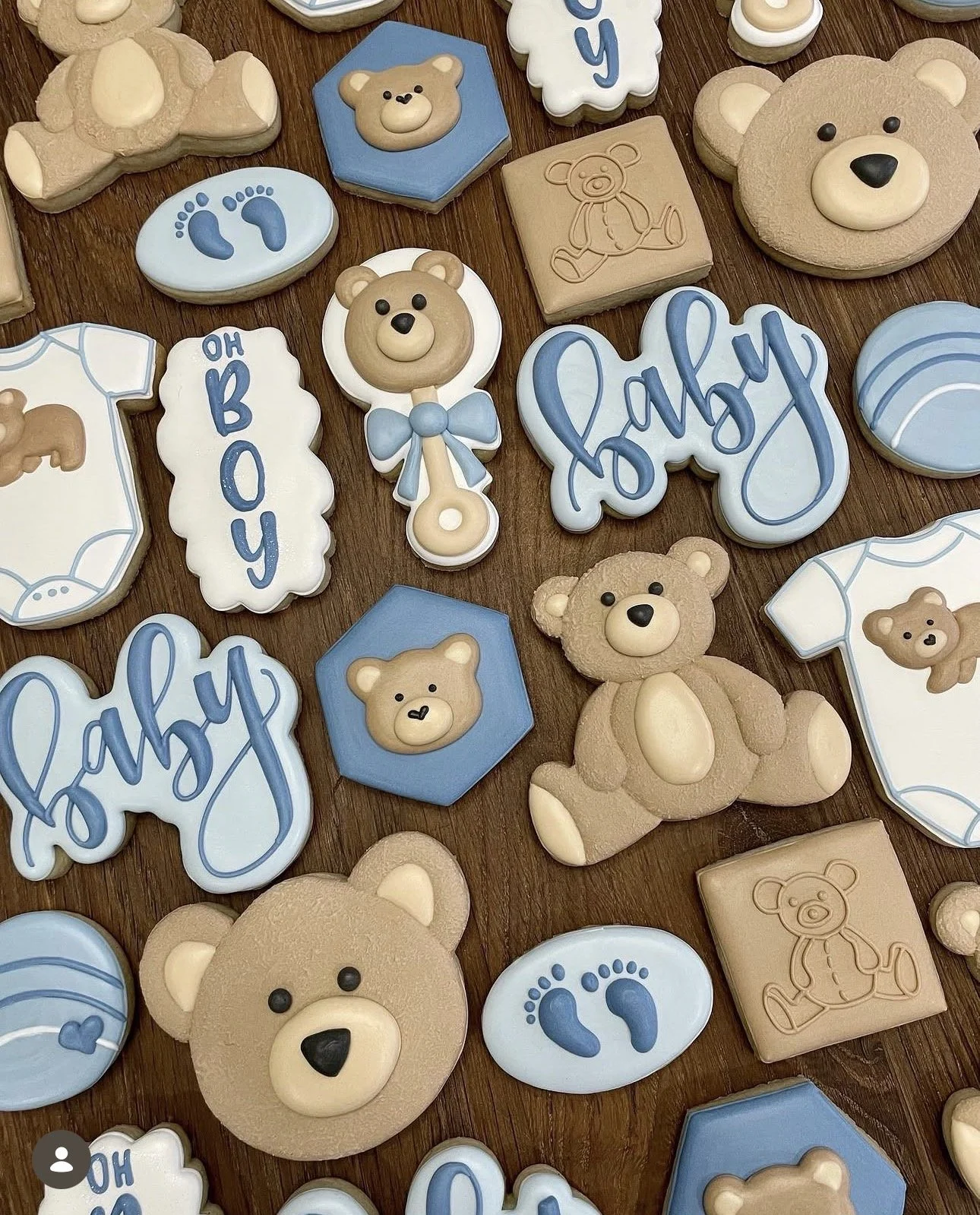 Decorated baby shower cookies featuring teddy bears, blue footprints, baby bottles, bibs, and the words “Baby Boy” on a wooden table.