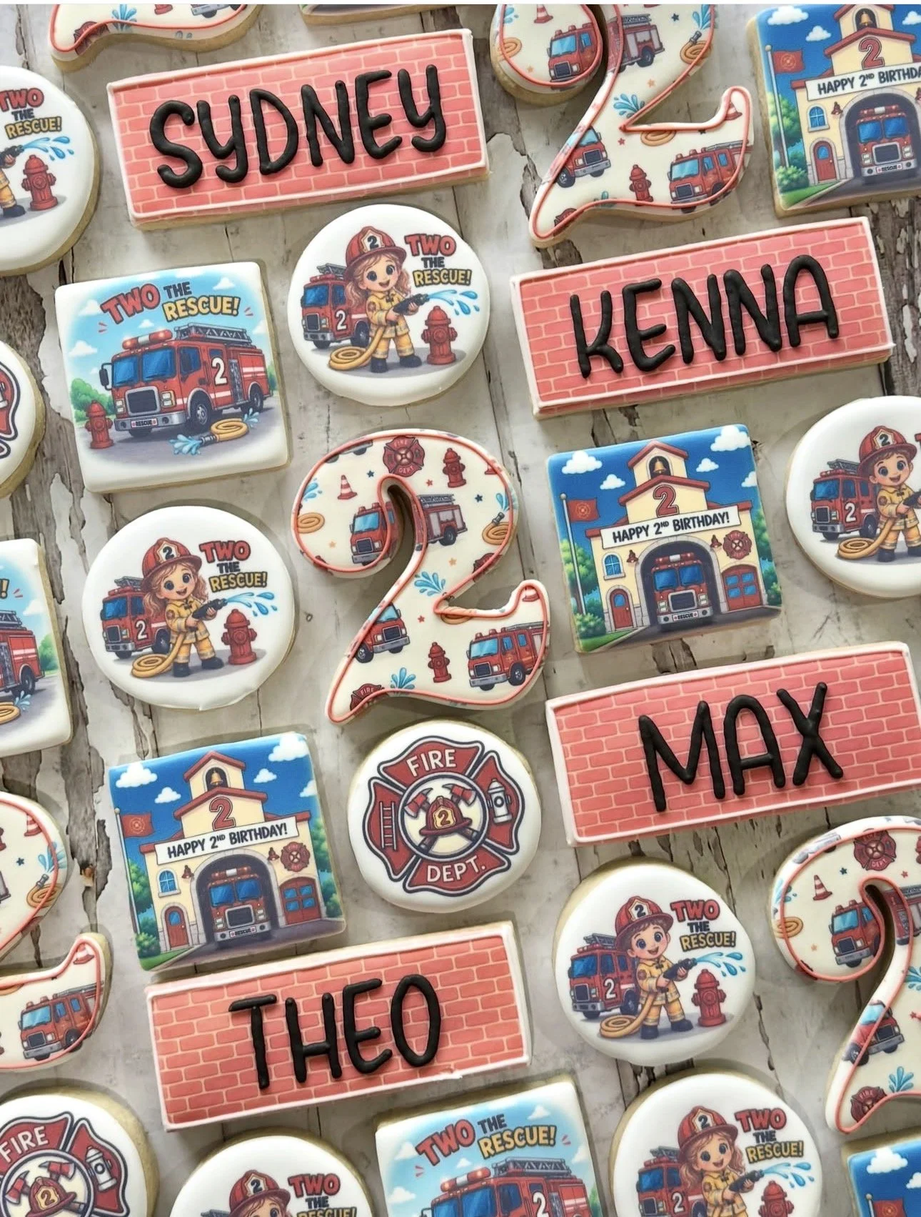 Assorted firefighter-themed second birthday cookies featuring fire trucks, firefighters, fire hydrants, and personalized names Sydney, Kenna, Max, and Theo.