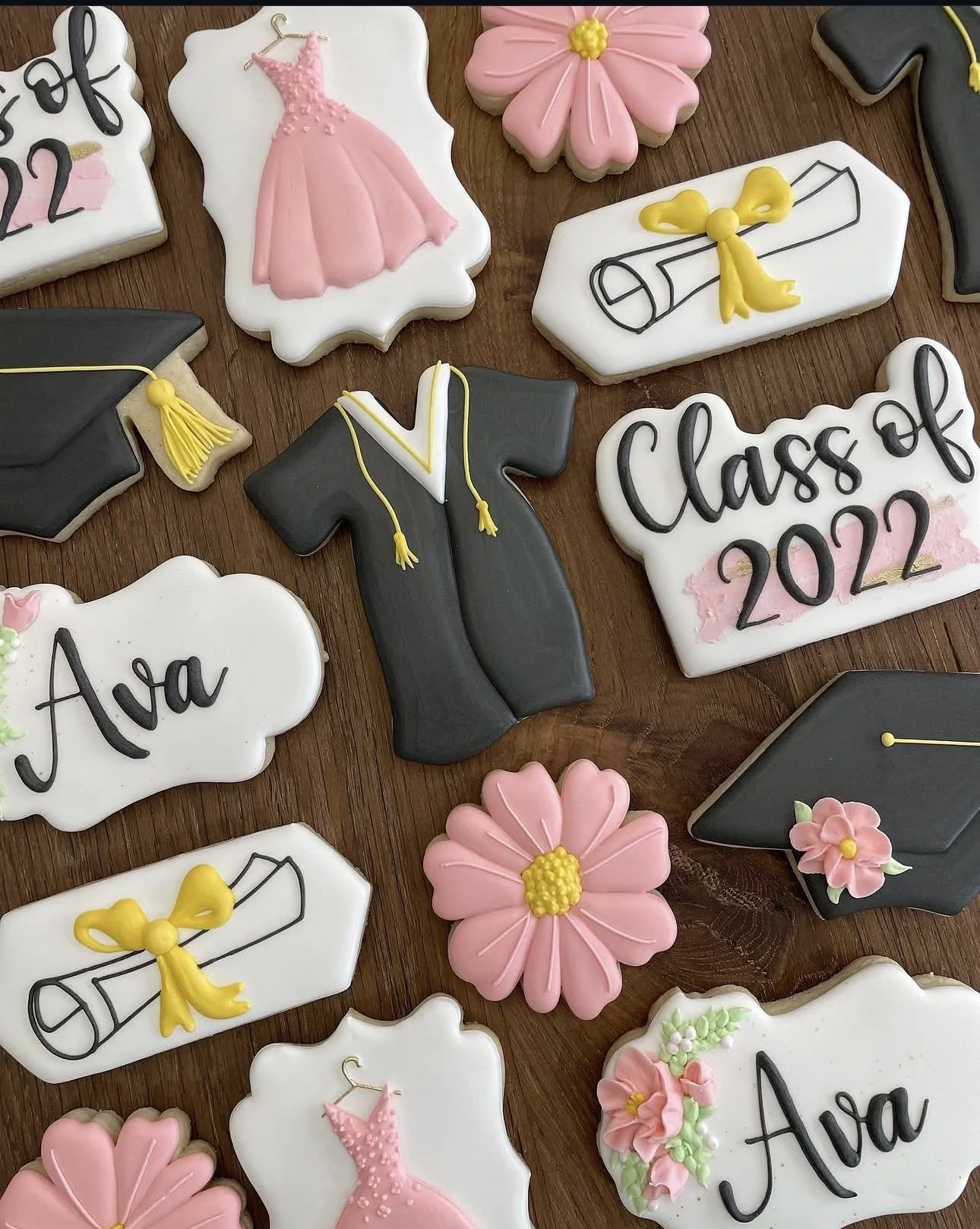 Graduation-themed decorated sugar cookies featuring black caps and gowns, pink dresses and flowers, diplomas with yellow ribbons, and personalized Class of 2022 cookies.