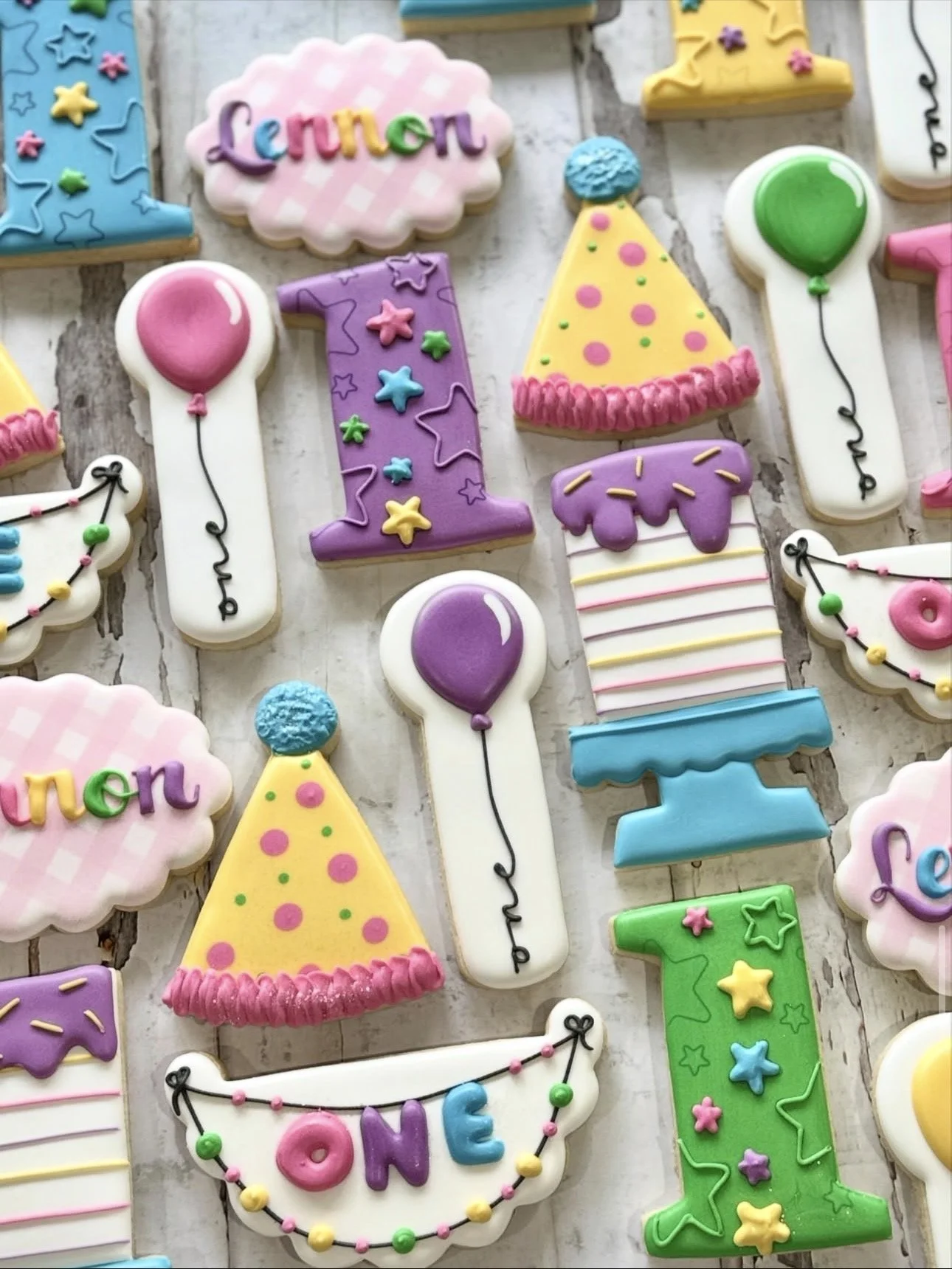 Colorful birthday-themed decorated sugar cookies with party hats, balloons, numbered candles, and “ONE” and “Lennon” lettering