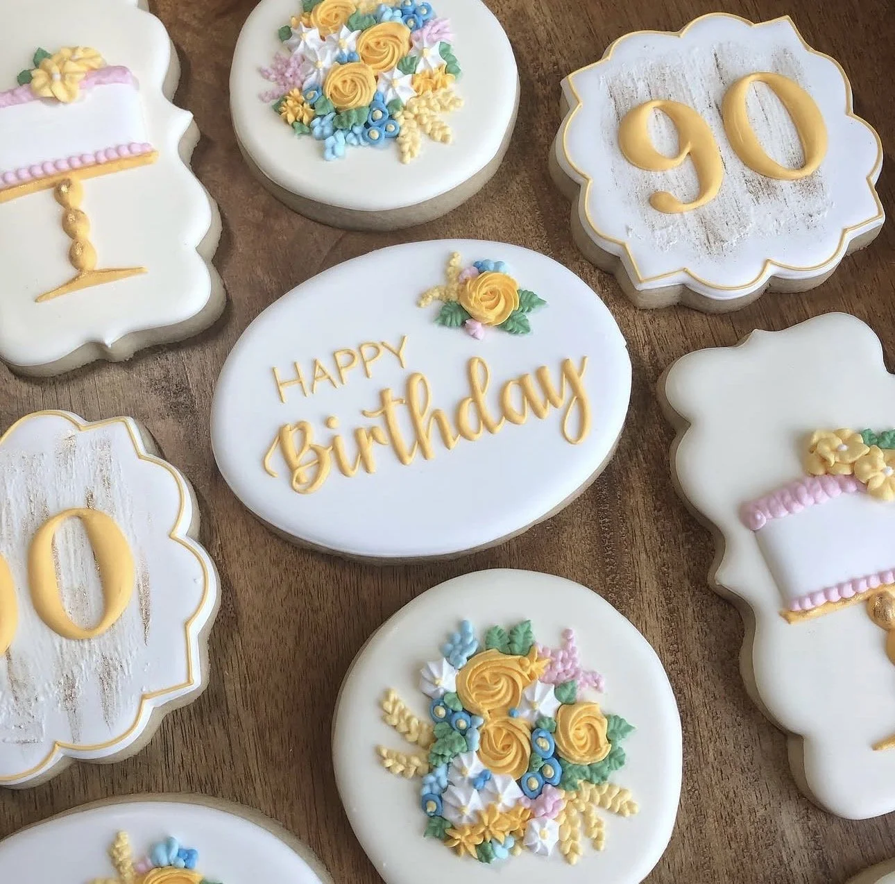 Decorated birthday cookies on a wooden table, including floral designs, the number 90, and a central cookie reading “Happy Birthday.”