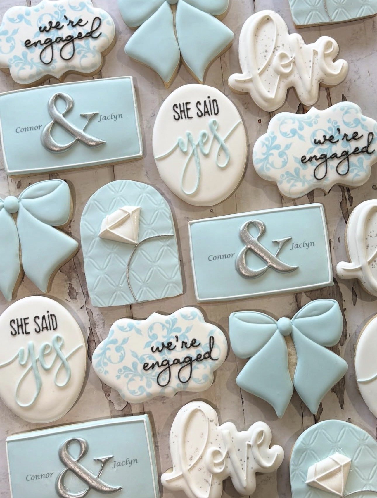 Blue and white engagement-themed decorated cookies featuring “we’re engaged,” “she said yes,” wedding bows, diamond rings, and Connor and Jaclyn name plaques.