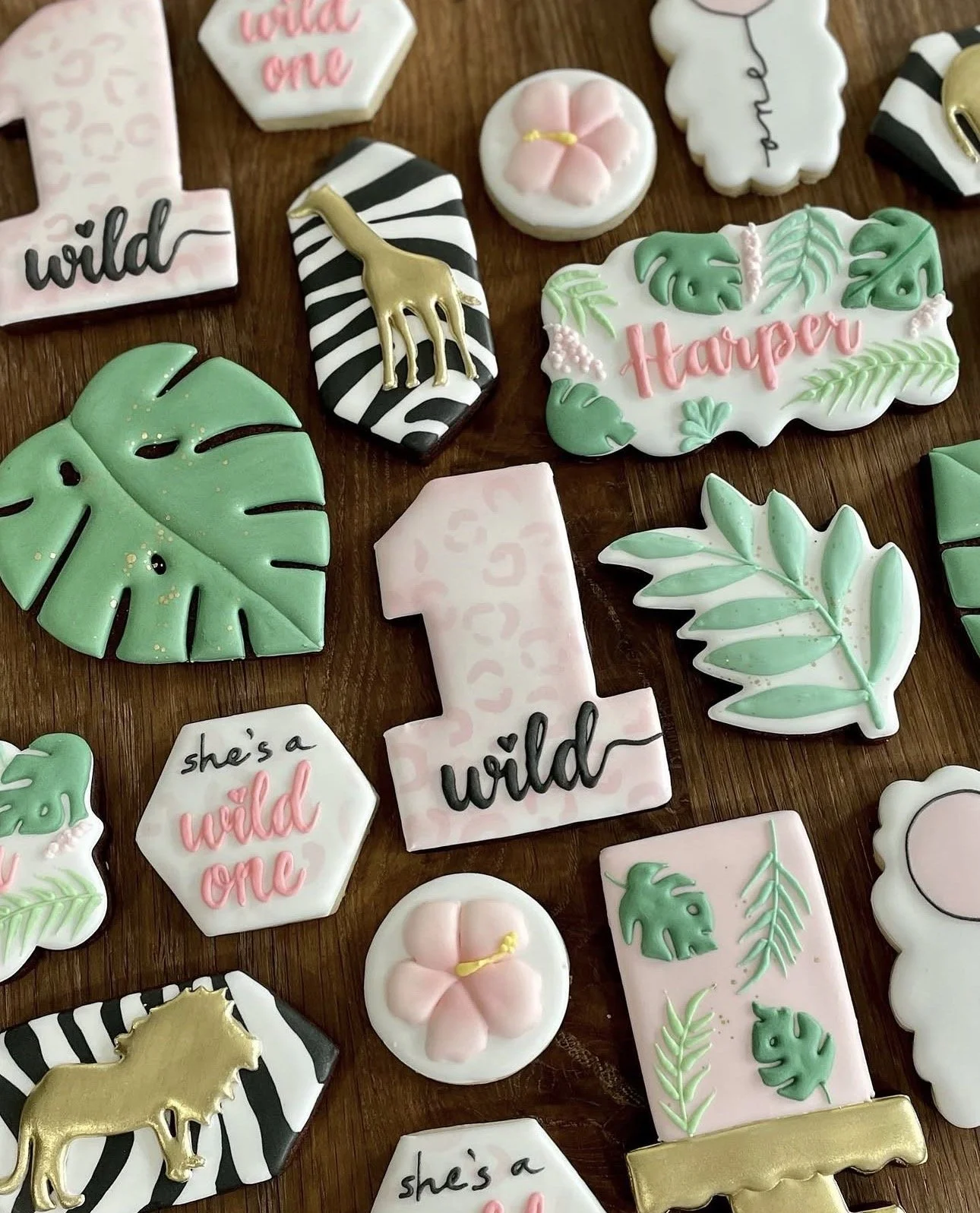 Decorated jungle-themed cookies featuring tropical leaves, pink flowers, zebra stripes, gold animal silhouettes, and “wild one” and “wild” messages.