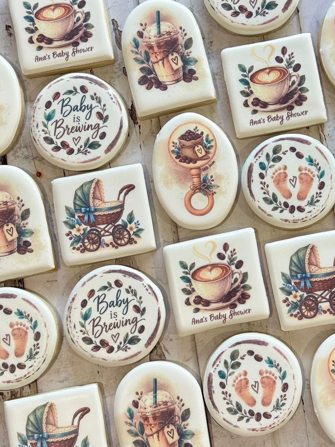 Assorted coffee-themed baby shower cookies reading “Baby is Brewing” and “Ana’s Baby Shower,” decorated with coffee cups, baby bottles, footprints, and a stroller.