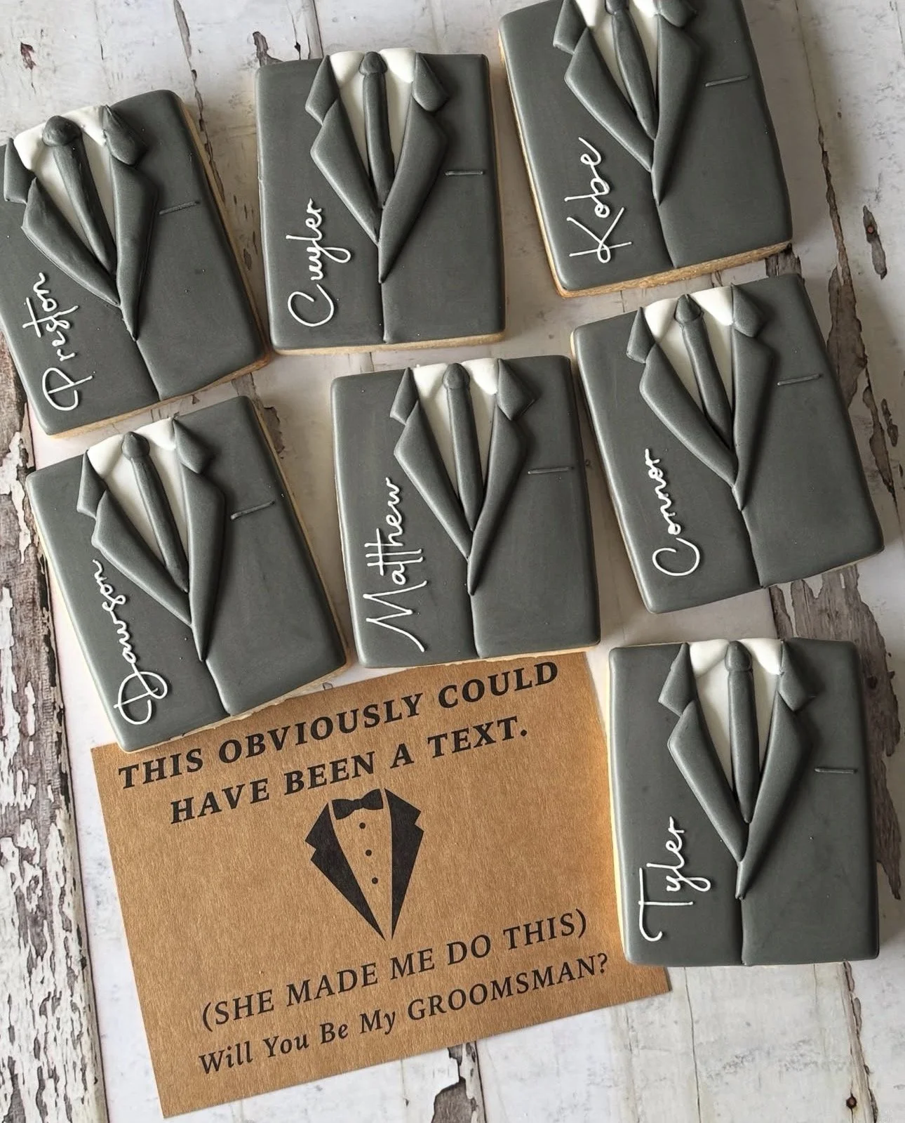 Handcrafted gray fondant tuxedo cookies personalized with groomsmen names, arranged around a card asking, “Will You Be My Groomsman?”