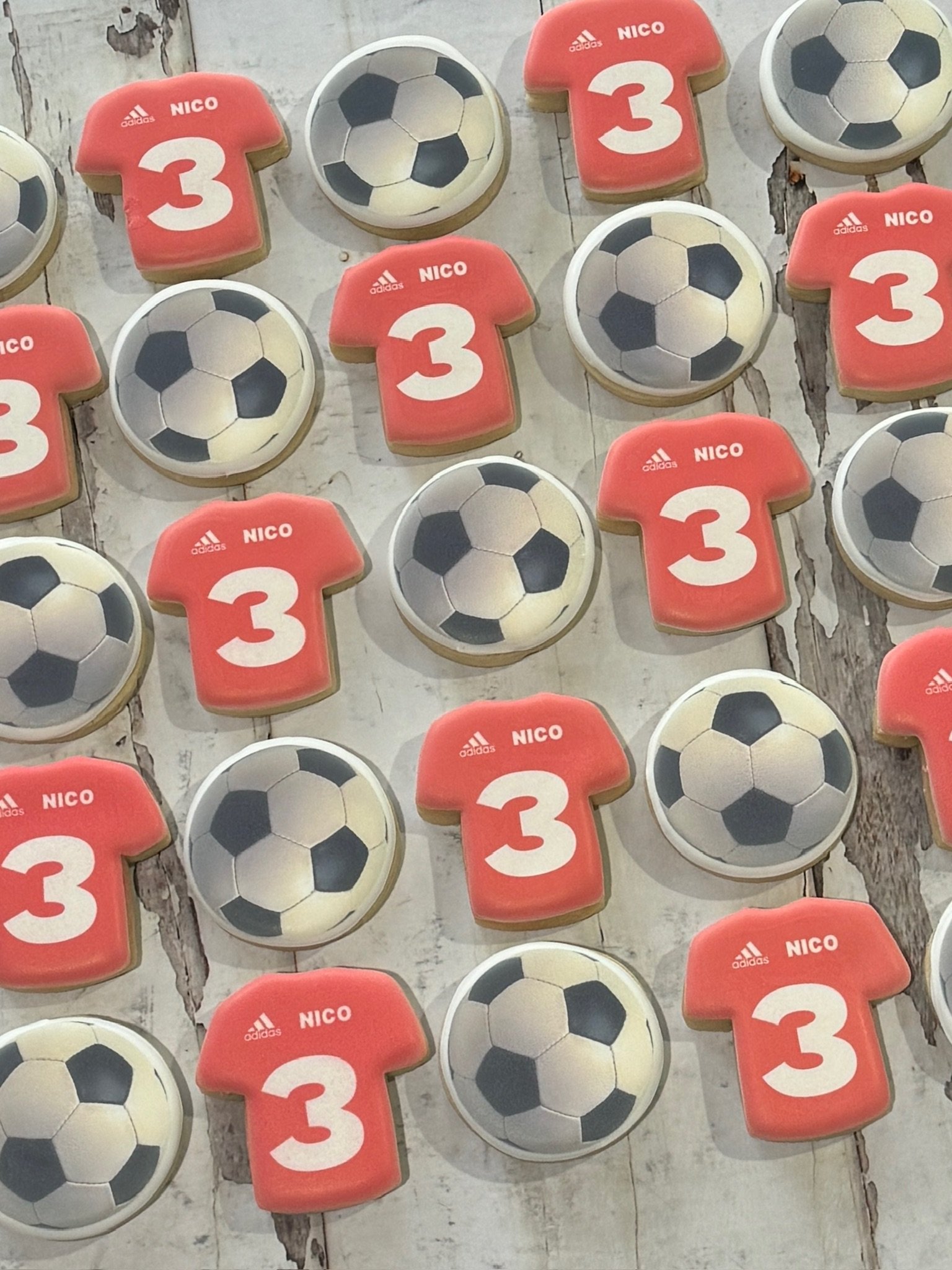Pattern of red soccer jersey-shaped cookies labeled “NICO” and number 3, alternating with black-and-white soccer ball cookies on a rustic wooden surface.