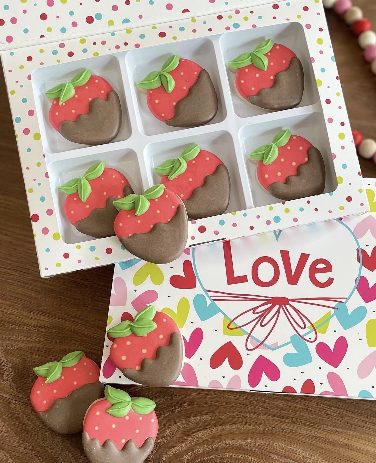 Strawberry-shaped chocolate-dipped cookies in a polka-dot gift box beside colorful heart-themed wrapping paper.