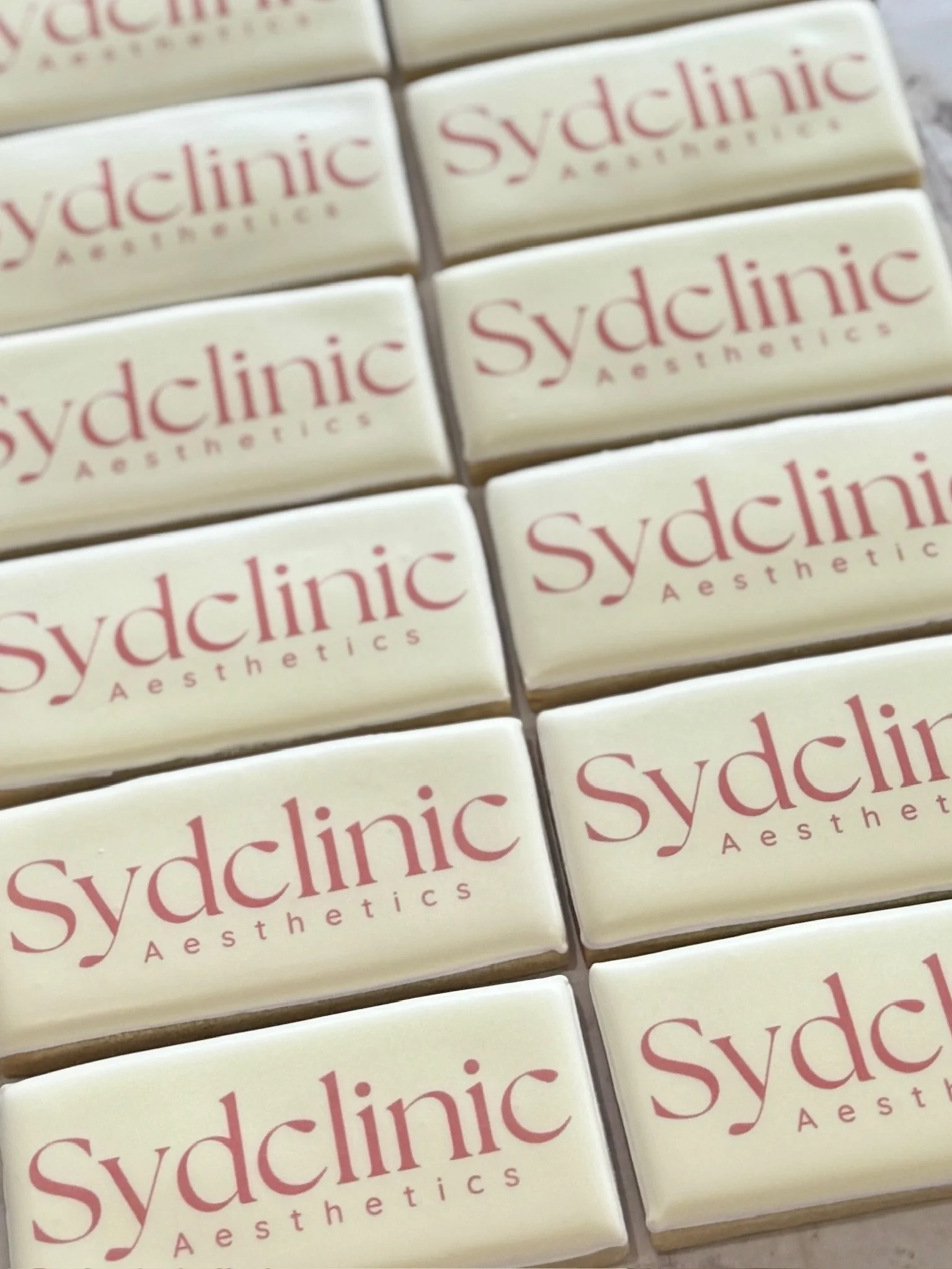 Close-up of cream-colored tiles arranged in a grid, each printed with the red “Sydclinic Aesthetics” logo.